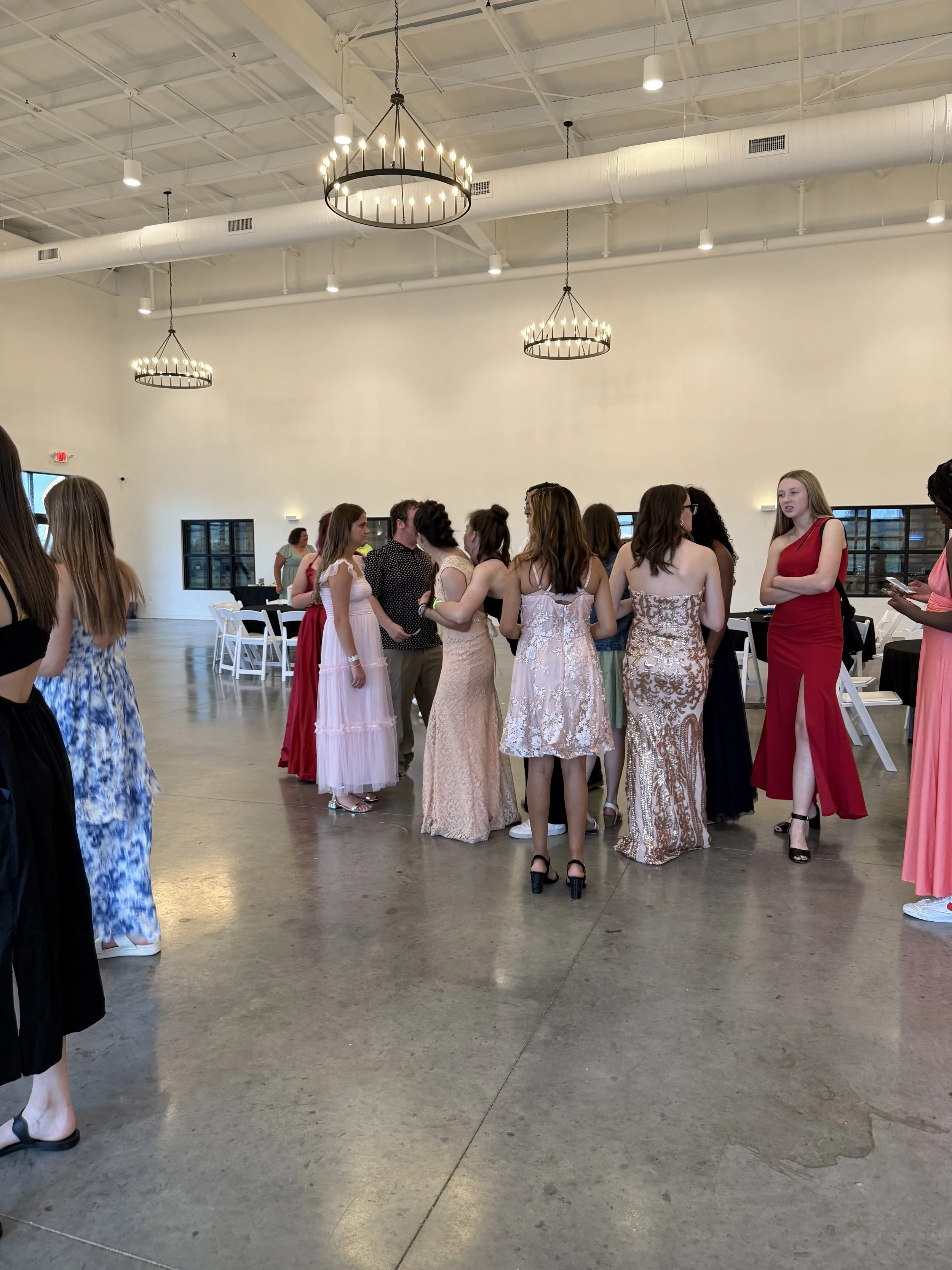 Group of women in formal dresses talking in a large, well-lit hall with modern chandeliers and a polished concrete floor.