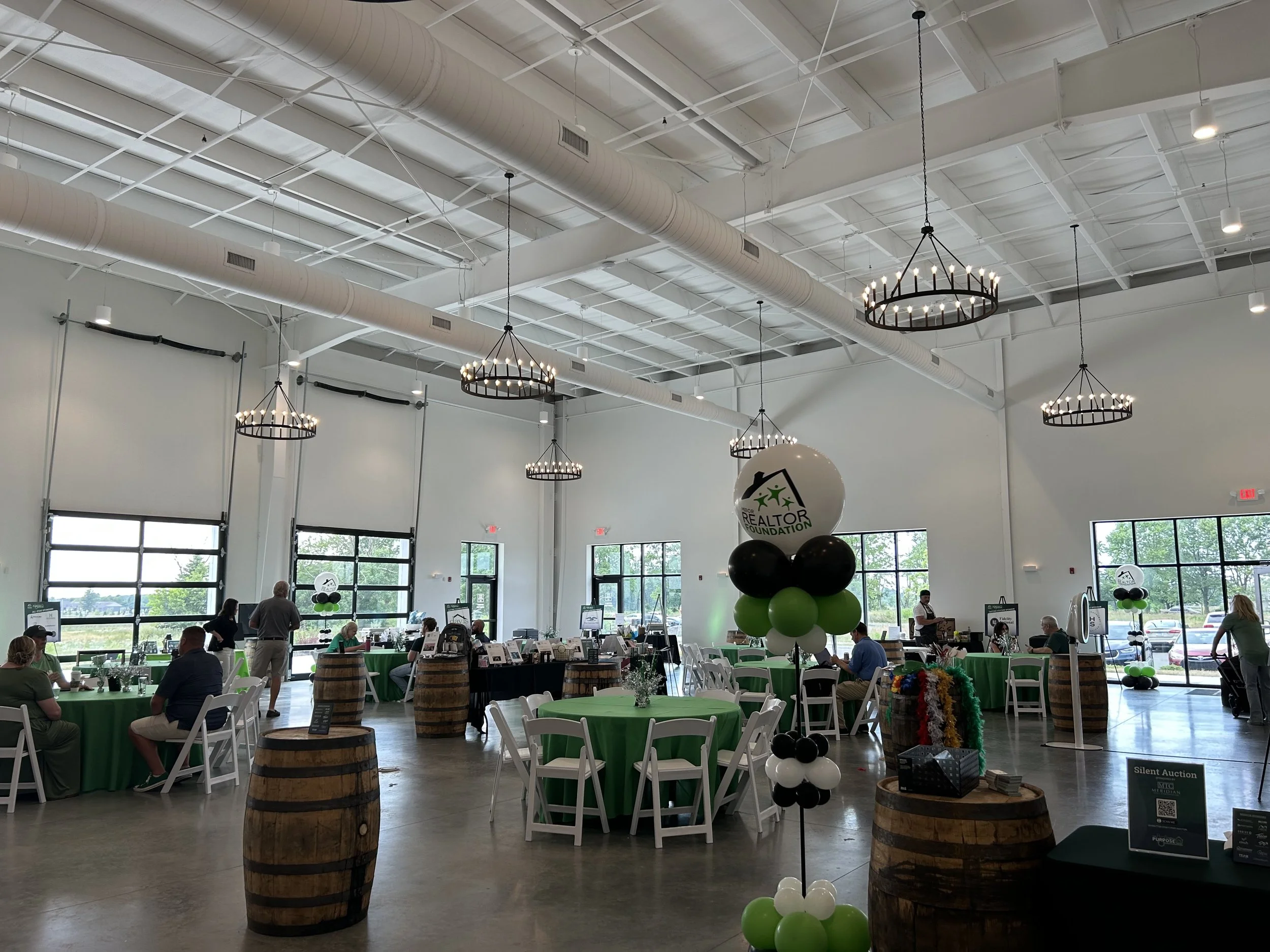 A spacious event hall decorated with green, black, and white balloons, with tables covered in green tablecloths and surrounded by white chairs. People are seated and standing, engaging in activities. The hall has large windows allowing natural light,