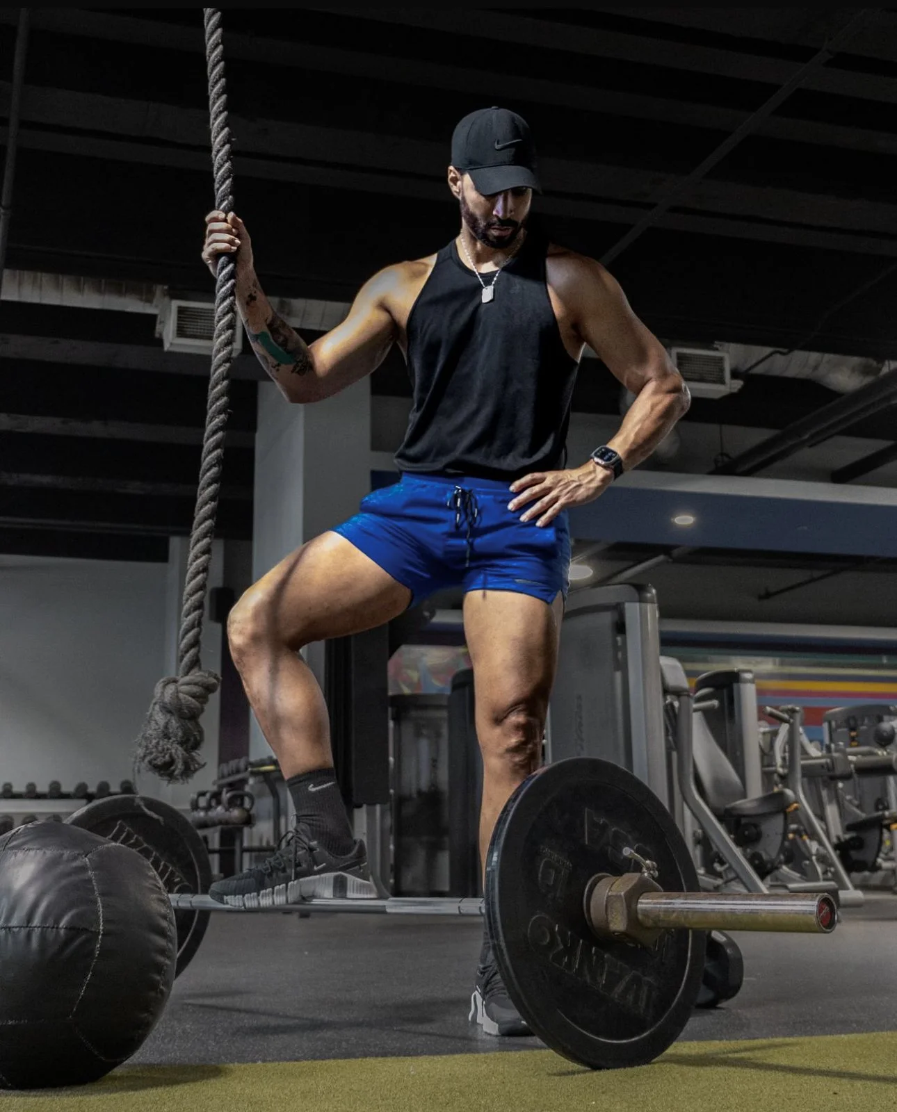 A muscular man in a black tank top, blue shorts, and a black cap working out with a barbell in a gym, holding a thick rope.