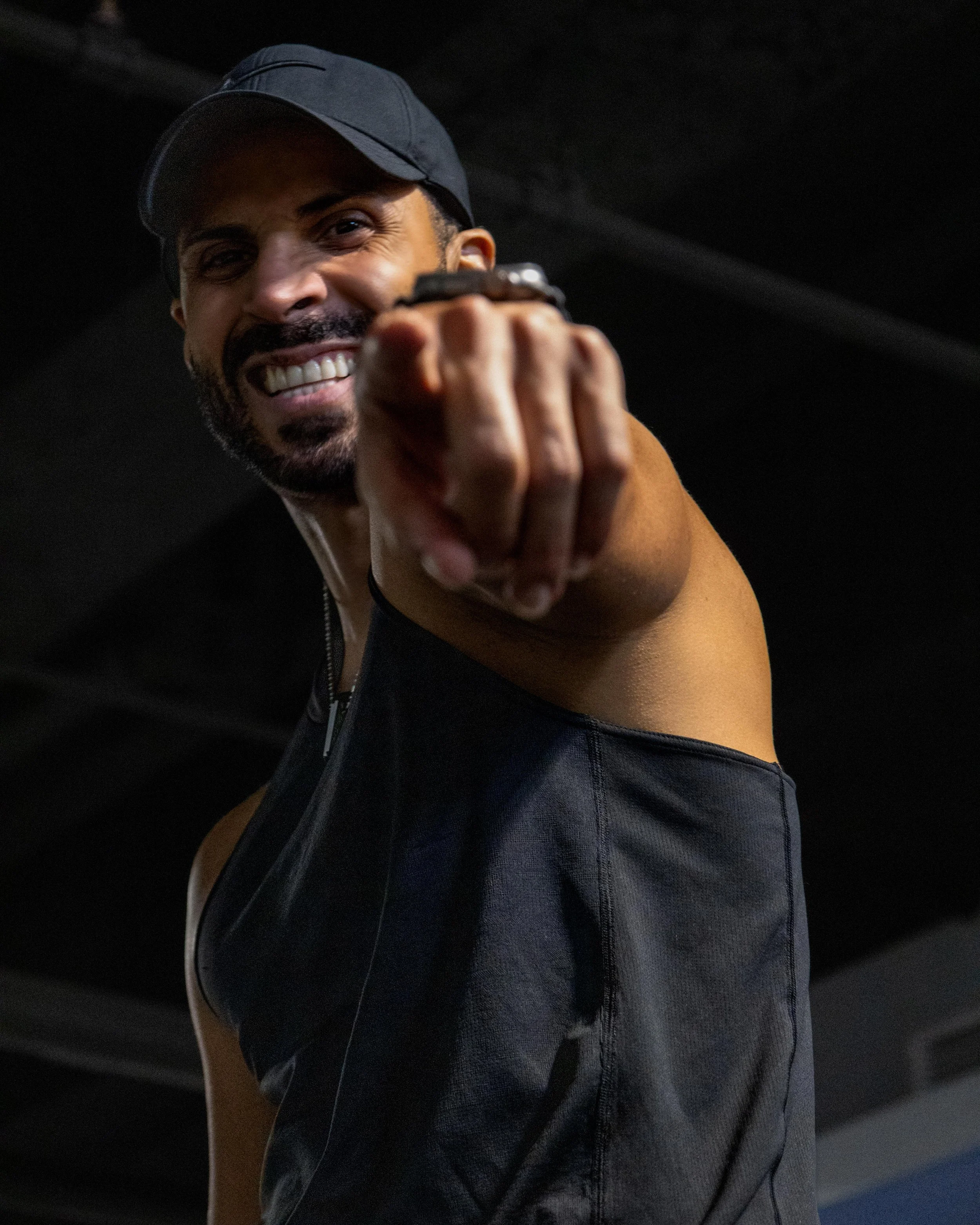 A man in a black cap and sleeveless athletic top smiling and pointing directly at the camera.