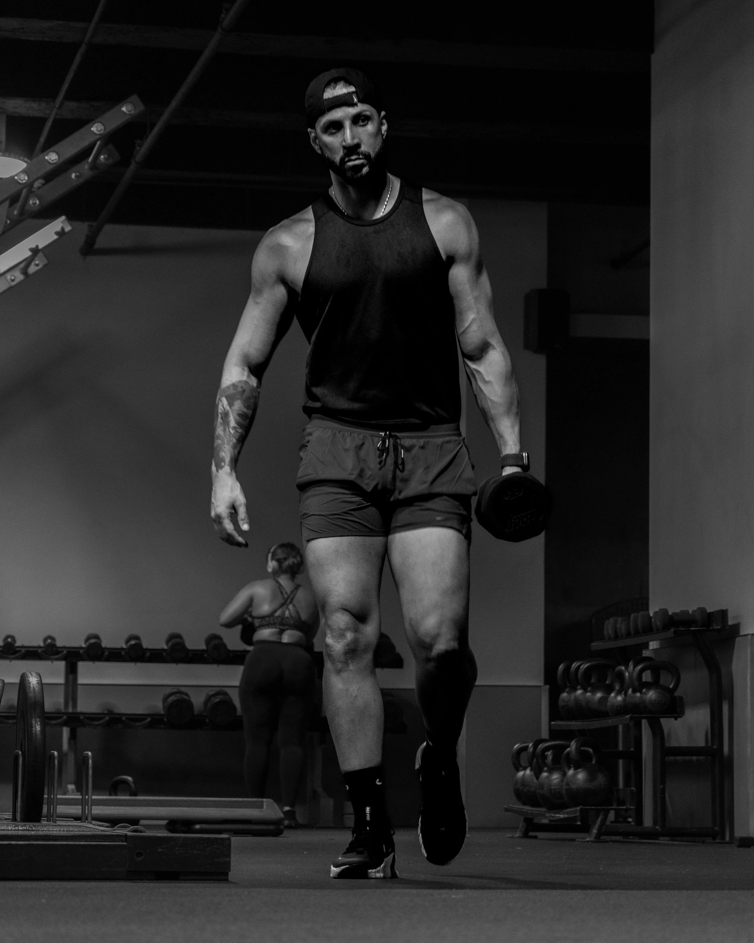 A man lifting a dumbbell in a gym, wearing a black sleeveless shirt, shorts, and a backwards cap, with another person in the background working out.