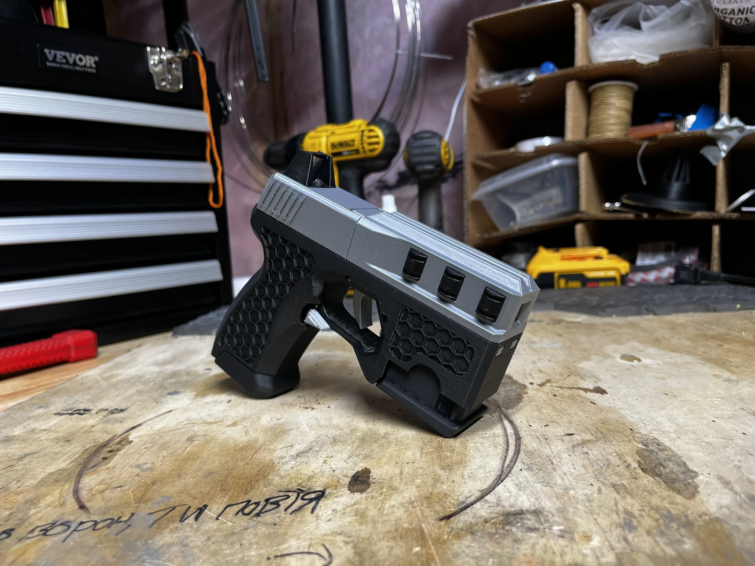 A small, 3D-printed coilgun model on a workbench in a workshop, with tools and storage compartments in the background.