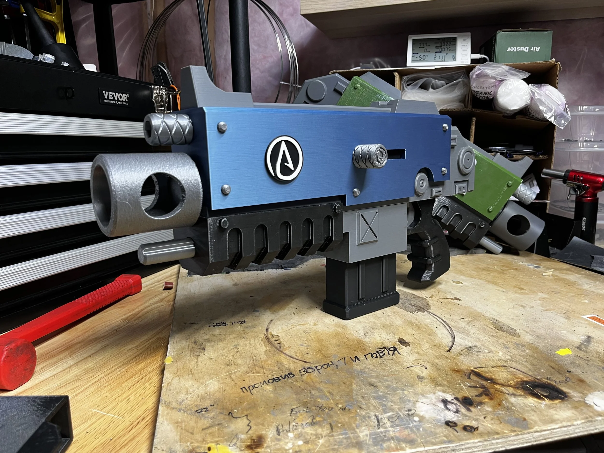 A custom-made prop gun resembling a sci-fi weapon is on a workbench in a workshop. It has a blue body, silver barrel, and various black and gray components, with metallic screws and textured details.