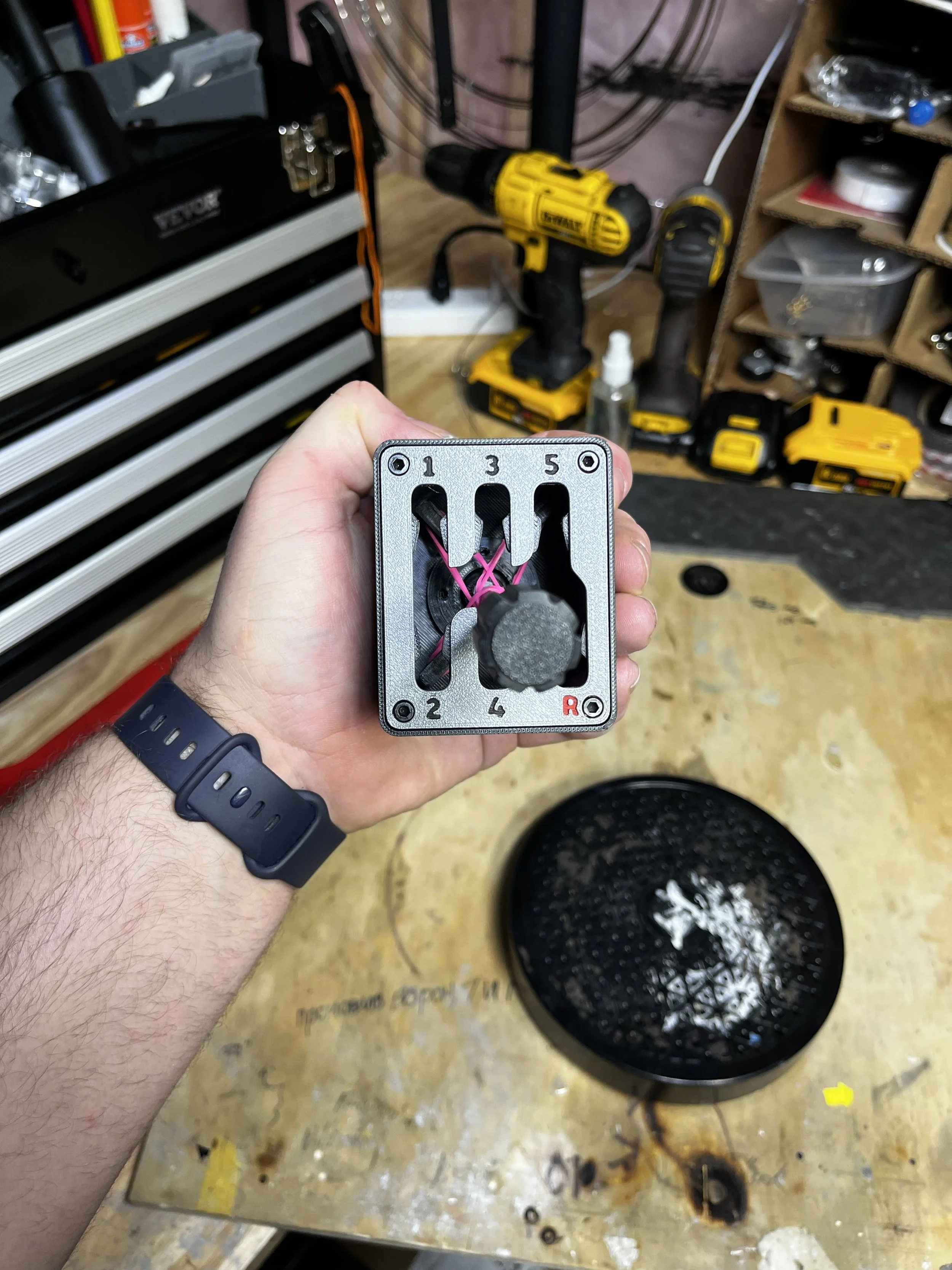 A person's hand holding a mechanical gear shifter plate with labeled gear positions (1, 2, 3, 4, 5, R) in a workshop or garage. There are tools, a yellow drill, and tool storage in the background.