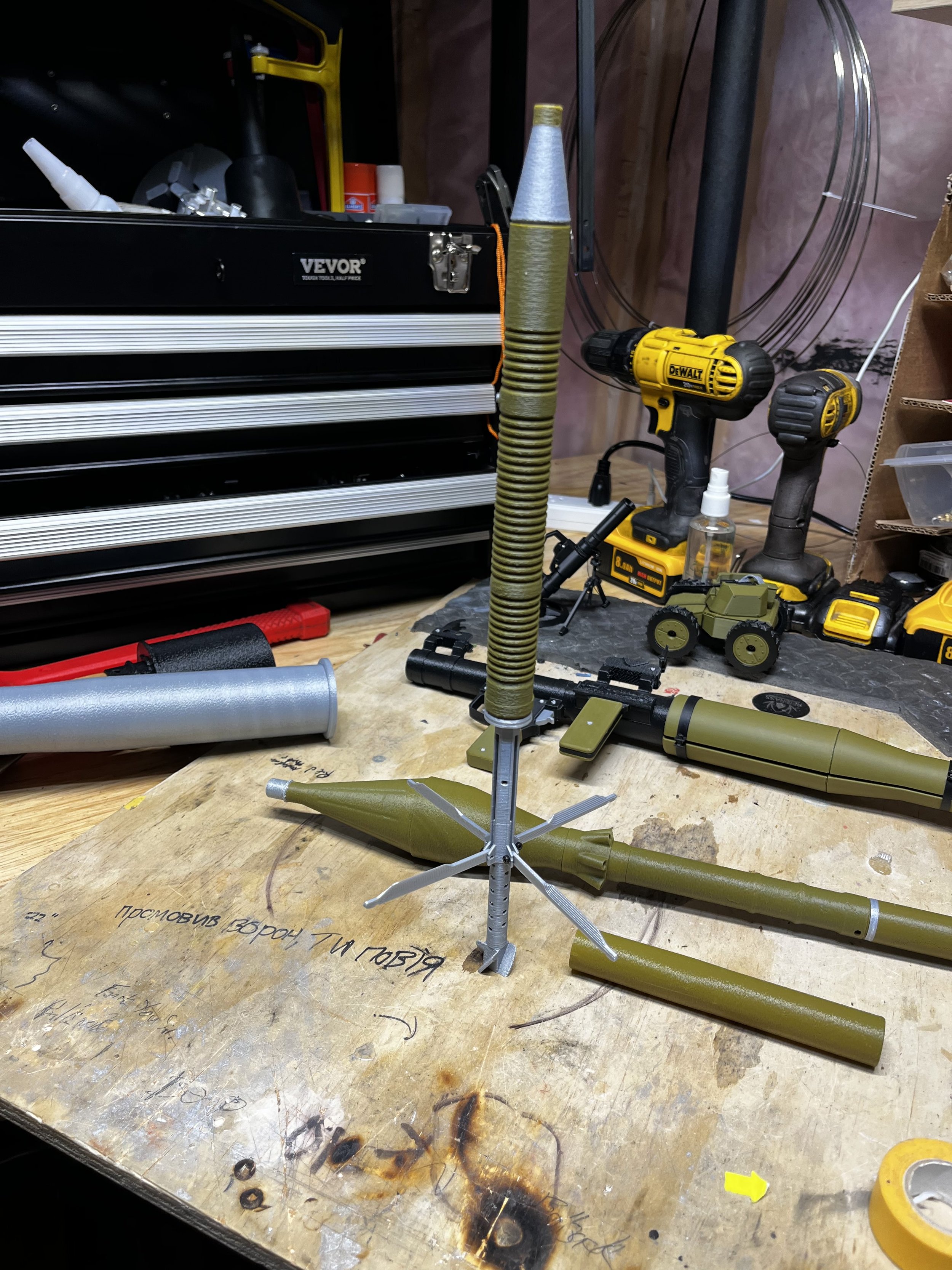 A workbench with parts and tools for assembling a model missile, including a large missile body, a nozzle, a circular base, and other components, with power tools and storage drawers in the background.