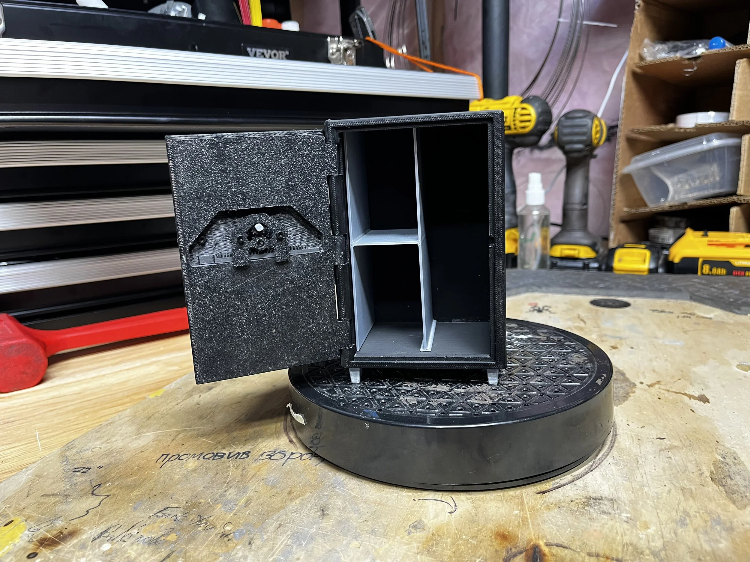 Small black safe with an open door, placed on a circular stand on a wooden workbench in a workshop.