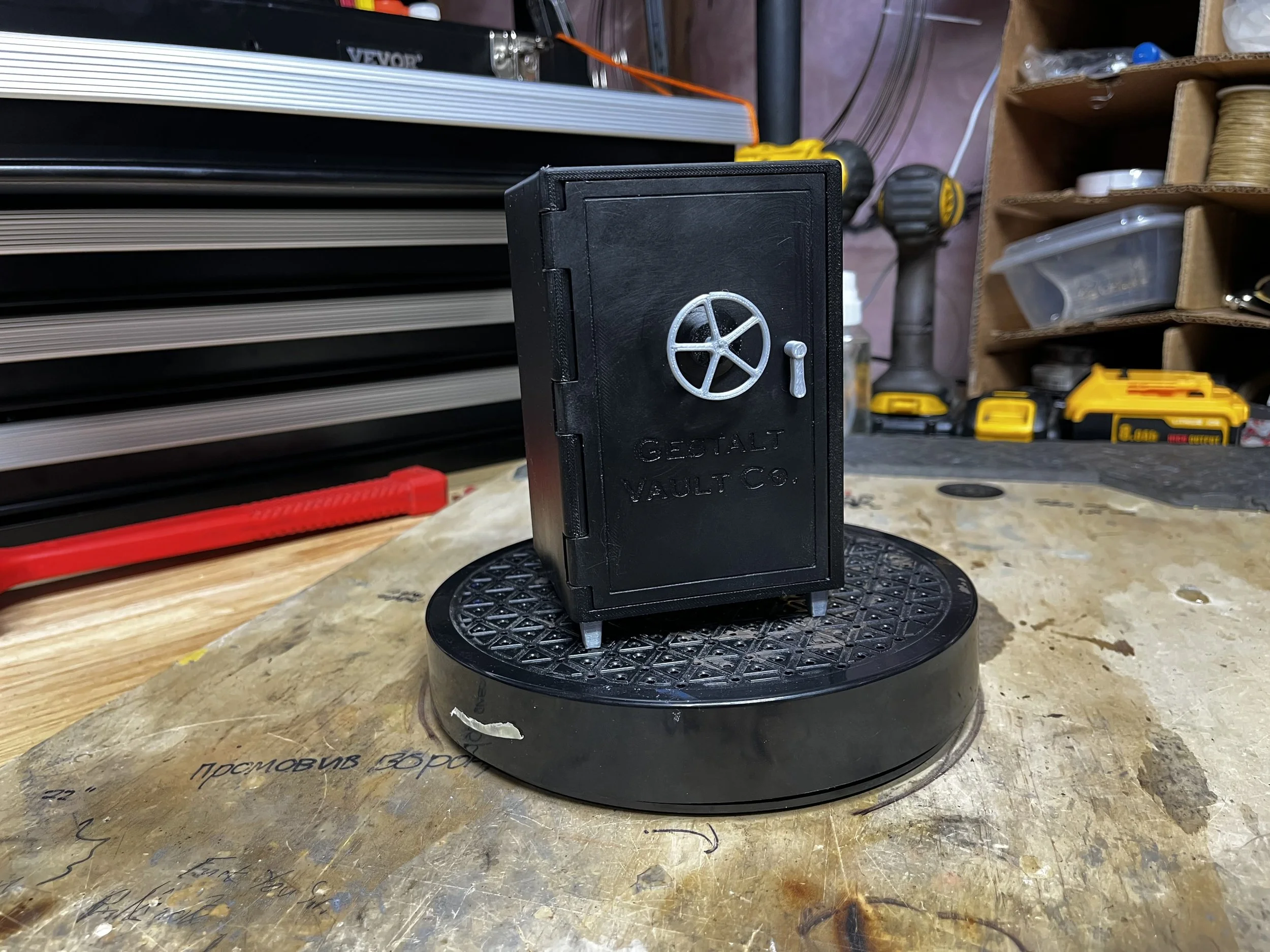 A black miniature vault with a gray wheel and handle, labeled 'Gestalt Vault Co.', sits atop a round black platform on a cluttered workshop workbench with tools and storage bins in the background.