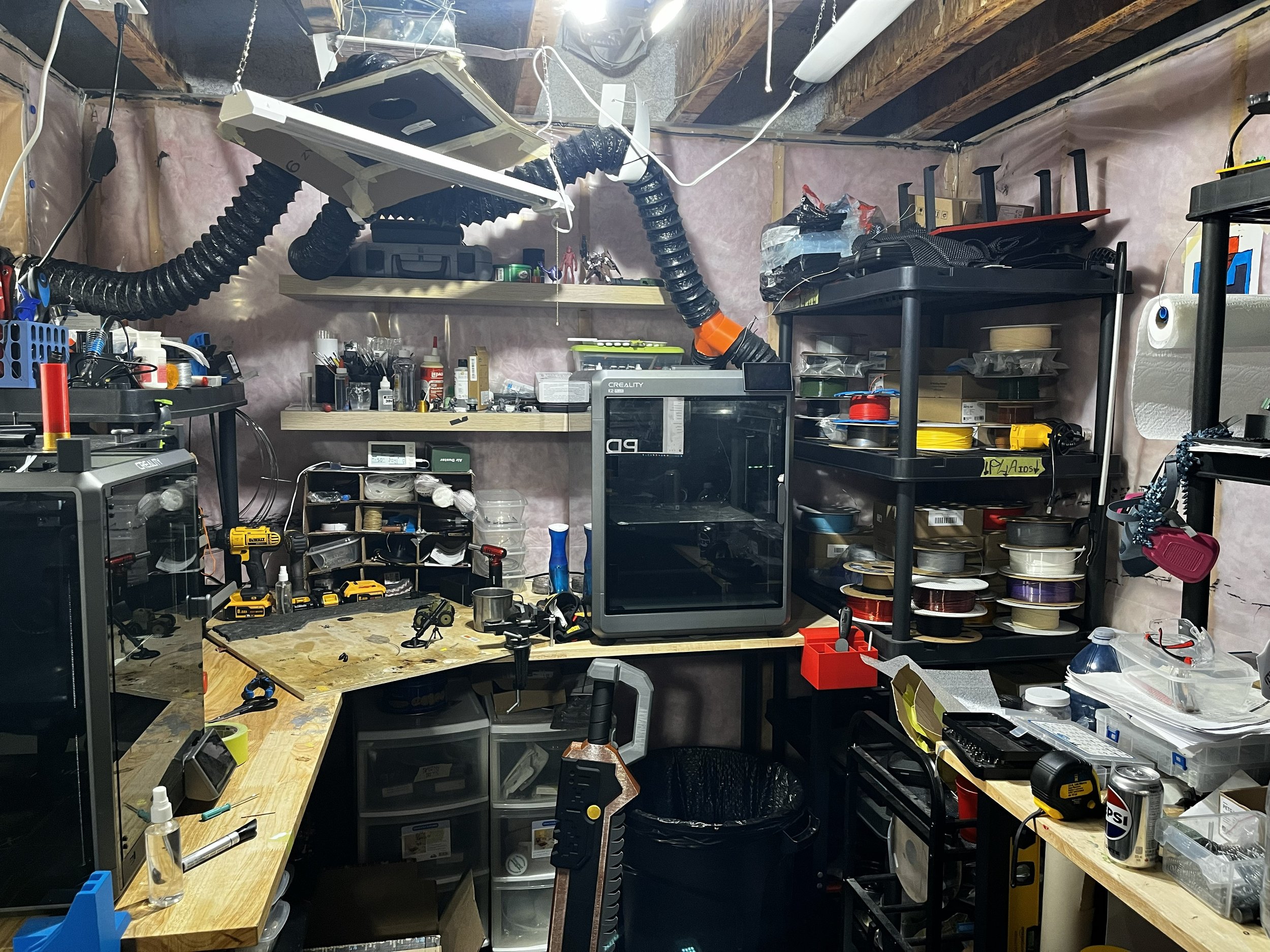 A cluttered workshop or garage workspace with shelves of tools, supplies, and equipment, including drills, containers, and electronic devices.