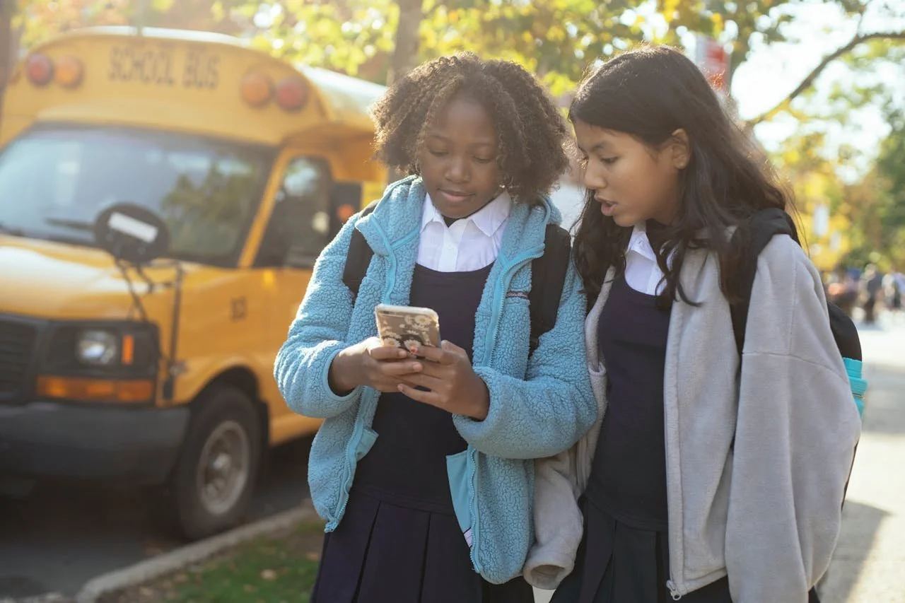 Two teenage girls in school uniforms standing by a yellow school bus, looking at a phone together, representing social media influence and external pressures on teen mental health in Alberta.