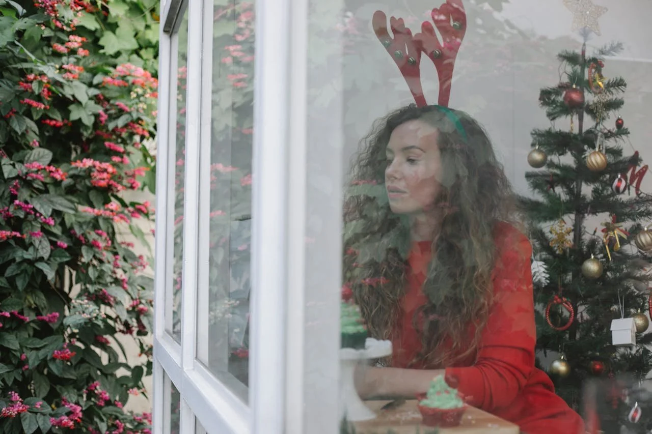 Woman standing quietly by a window beside a Christmas tree, reflecting during the winter holiday season.