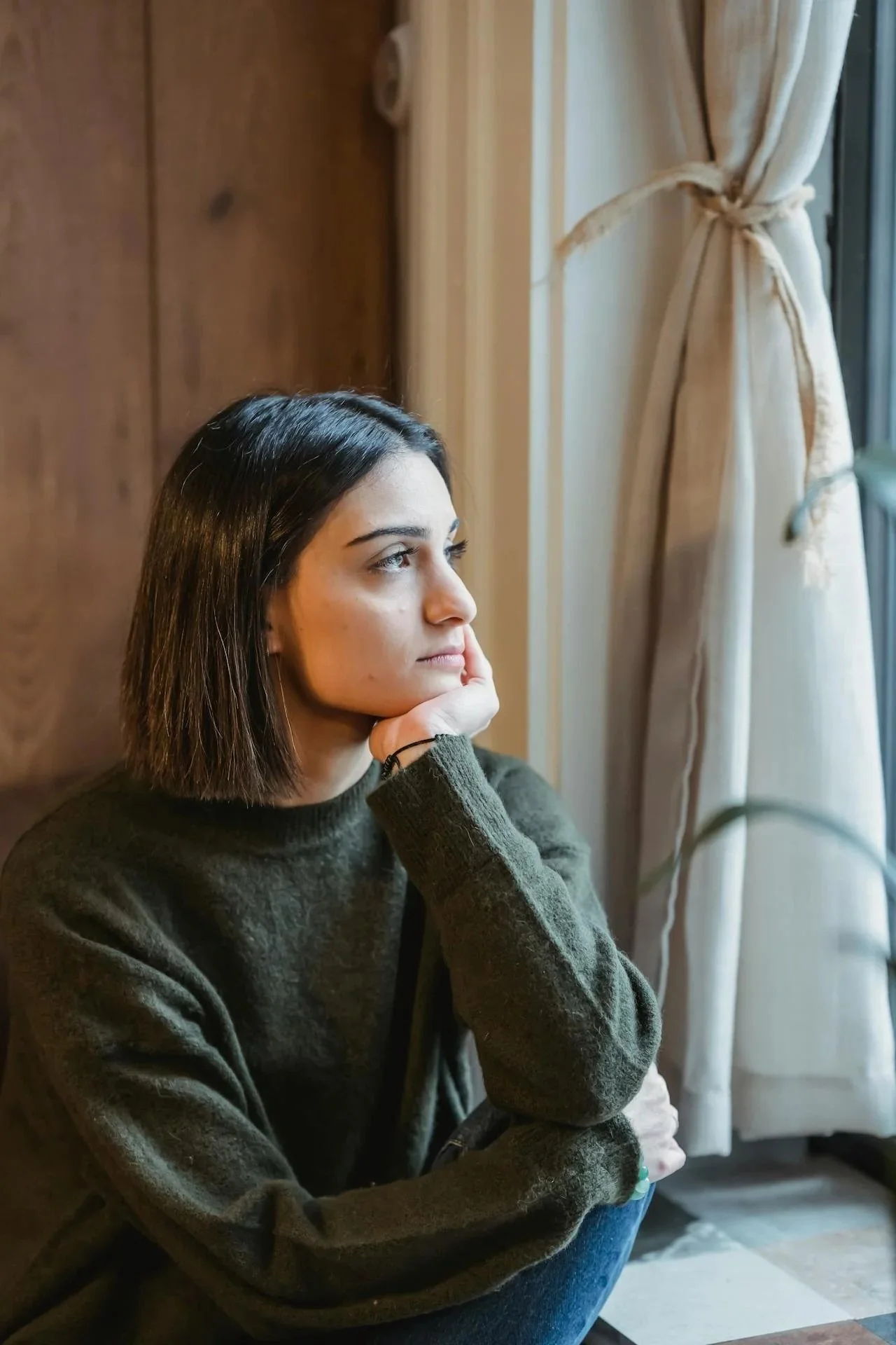 Woman sitting quietly by a window appearing emotionally distant while reflecting on the effects of past trauma