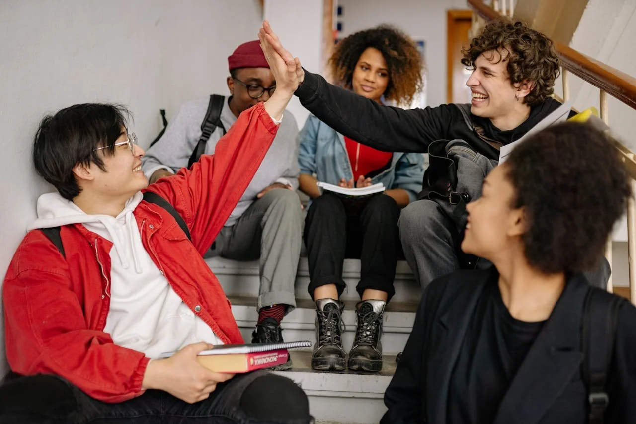 A diverse group of high school students sitting on stairs, smiling and high-fiving, representing peer support and connection in Alberta schools to promote student mental health.