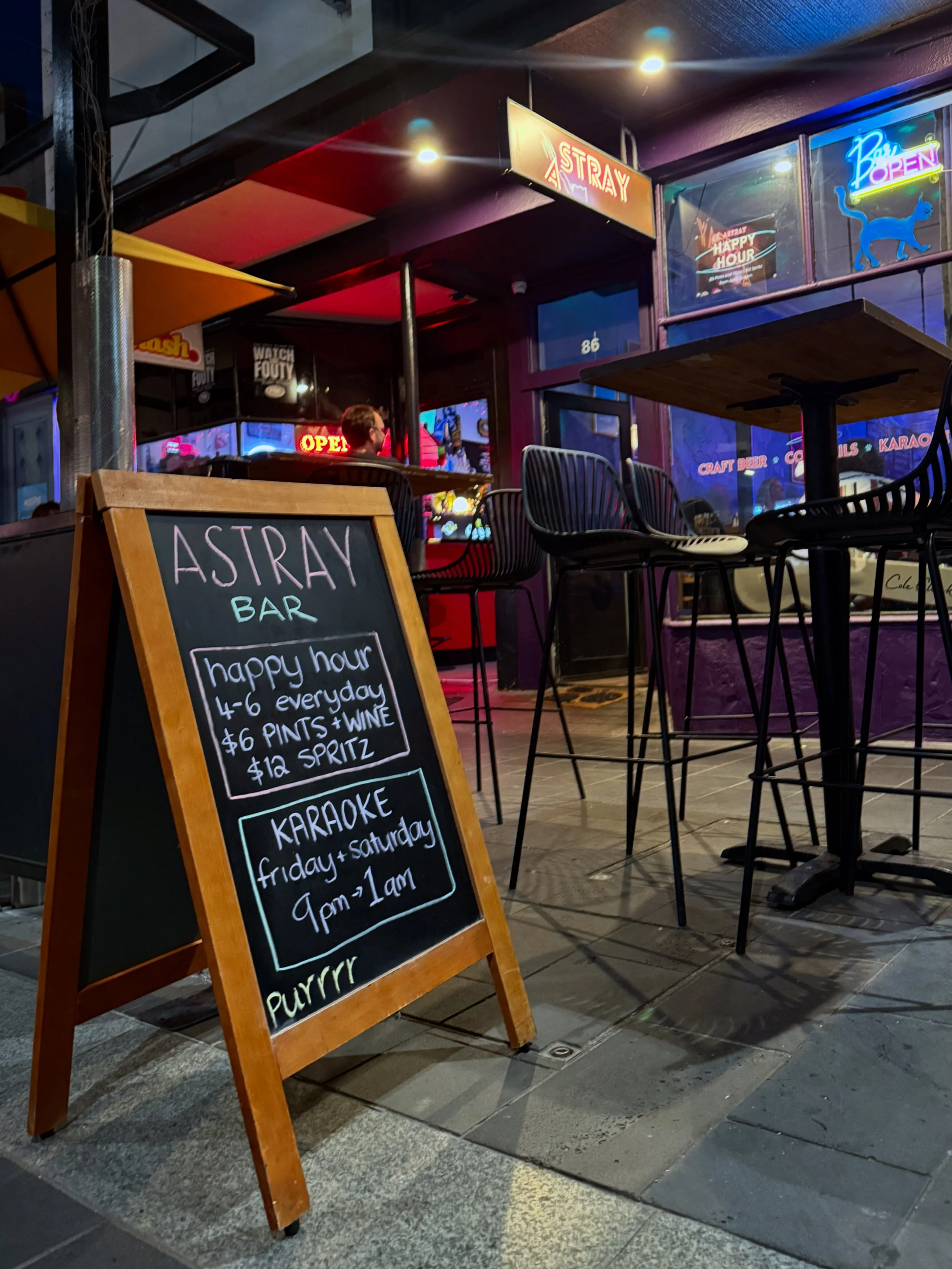 A blackboard sign outdoors advertising happy hour with $6 pints of house wine until 6 pm, next to two drinks on a wooden table; one is a yellowish beer and the other is a red cocktail with orange slices.