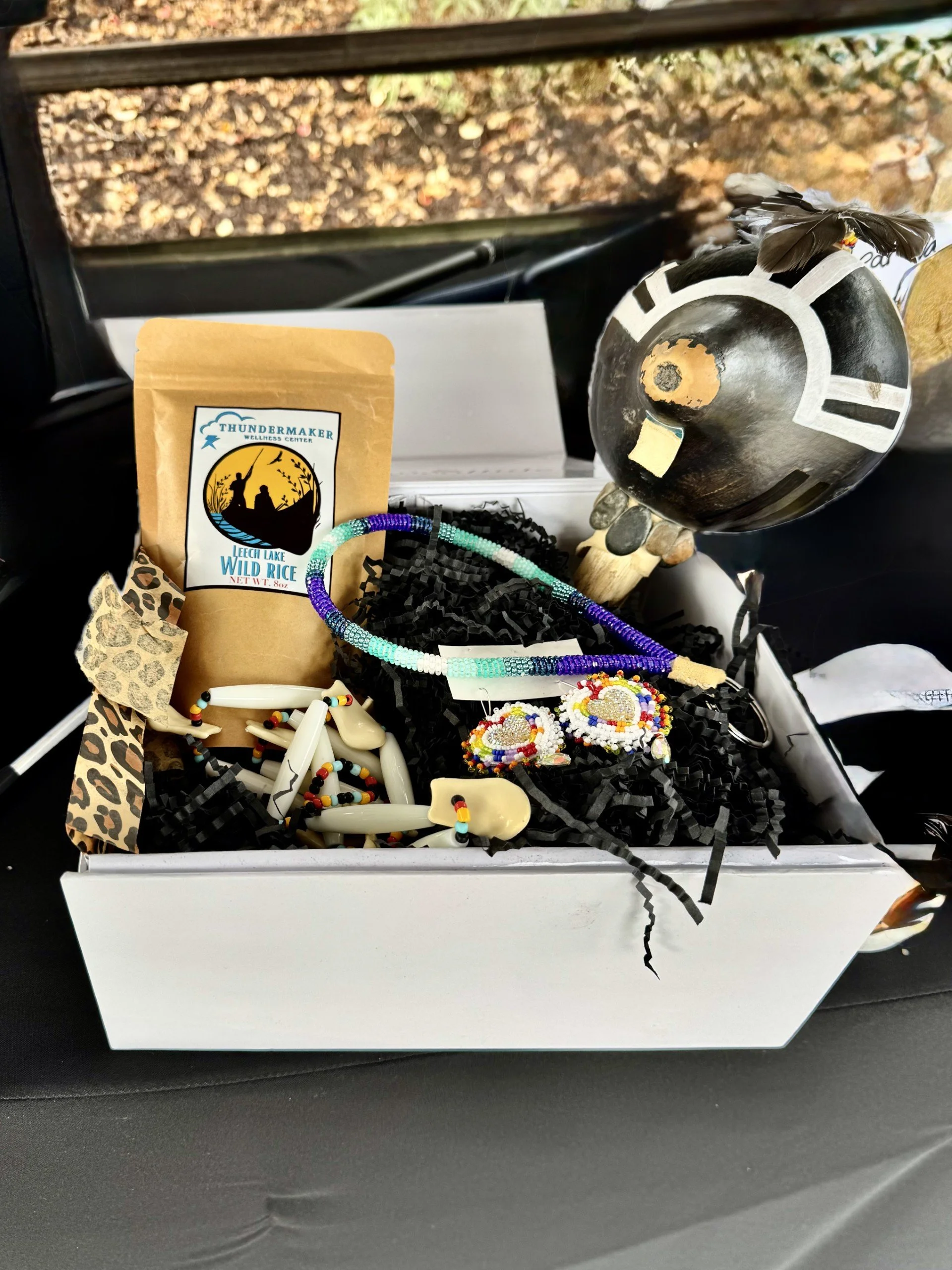 A white gift box containing handmade native american jewelry, a small bag of wild rice, and a decorative painted black and white gourd as a donation for a raffle item for the Native Youth Foundation Golf Tournament. 