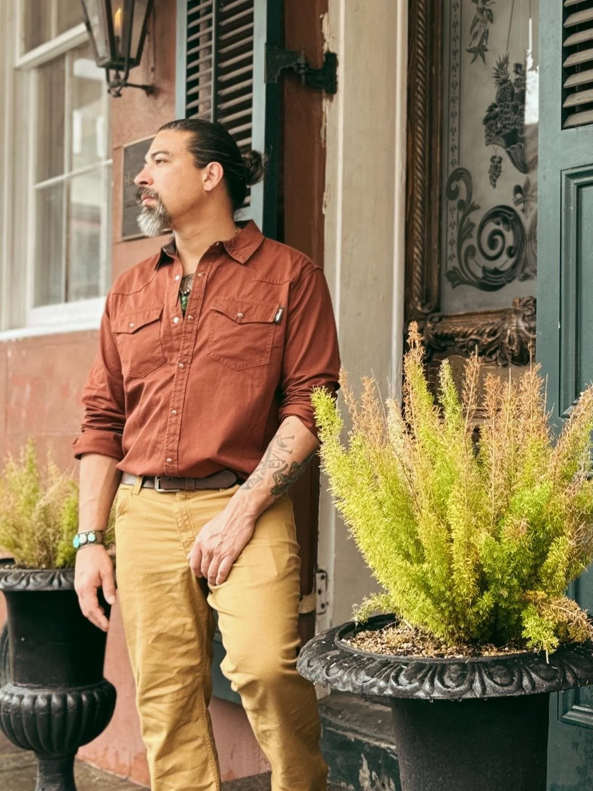 A man with long dark hair tied back, beard, and tattoos on his arm leaning against a building with potted plants nearby, wearing a brown button-up shirt and tan pants.