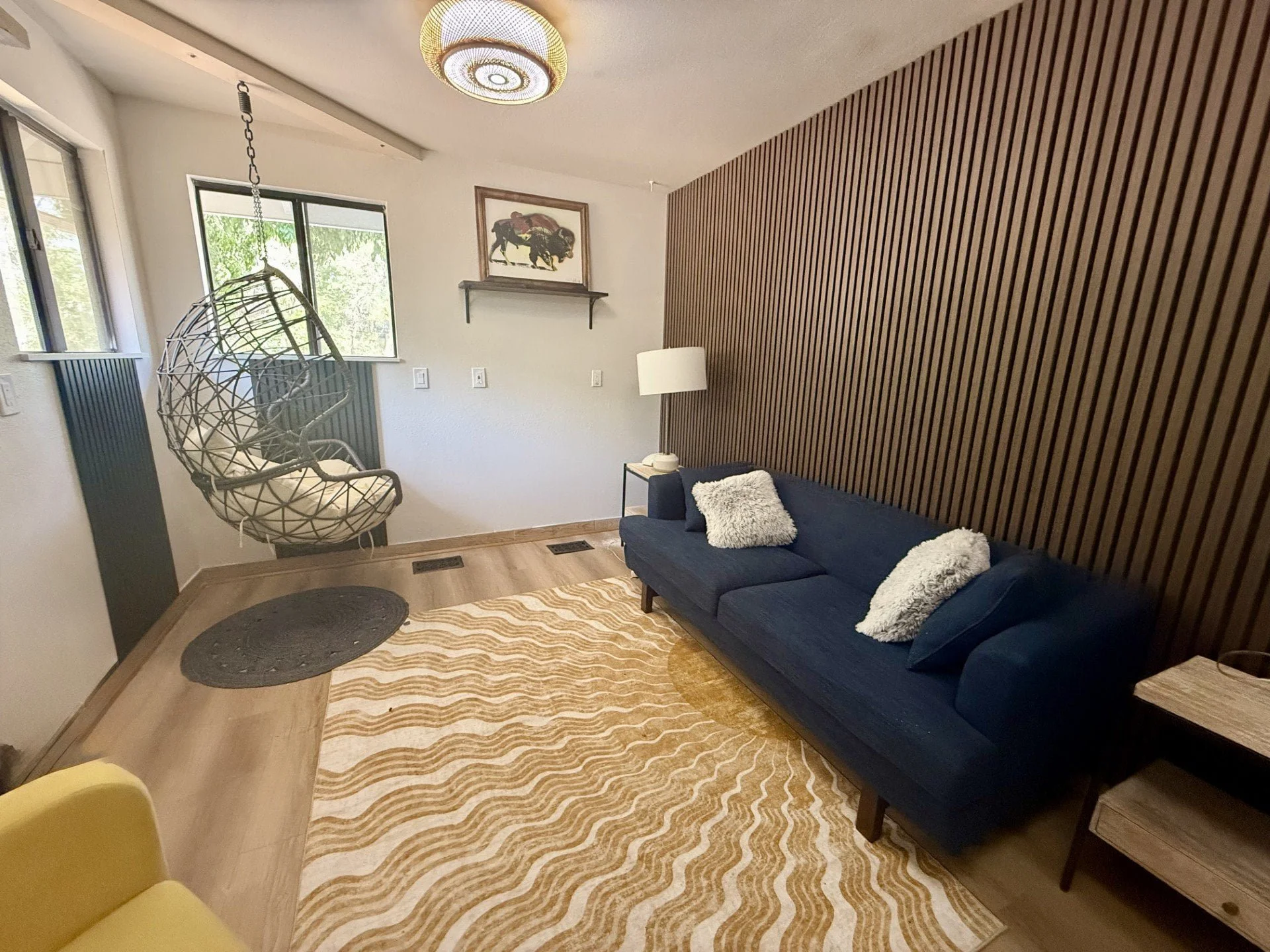A group therapy room or individual therapy room with a navy blue sofa, white fluffy pillows, wooden wall panel behind the sofa, hanging egg chair near window, white lamp on side table, and framed picture of a buffalo on the wall.