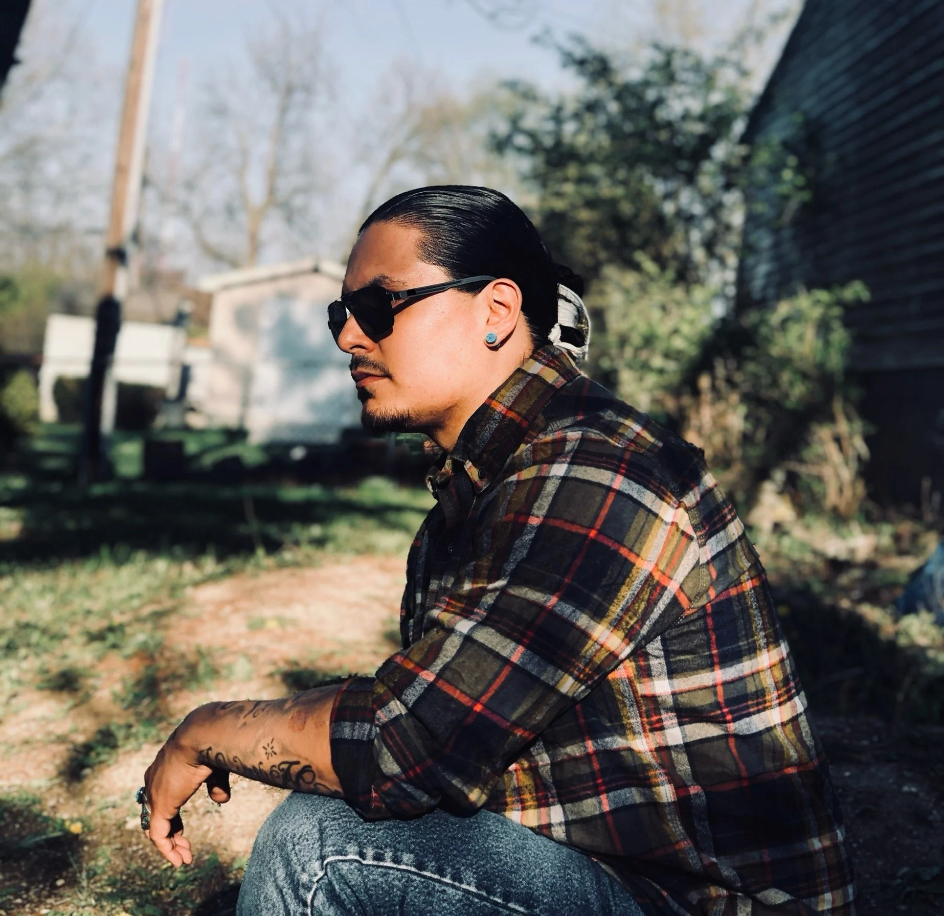 A man with long dark hair, sunglasses, and tattoos on his arm, wearing a plaid shirt, sitting outside on the ground in a yard with trees and a house in the background.