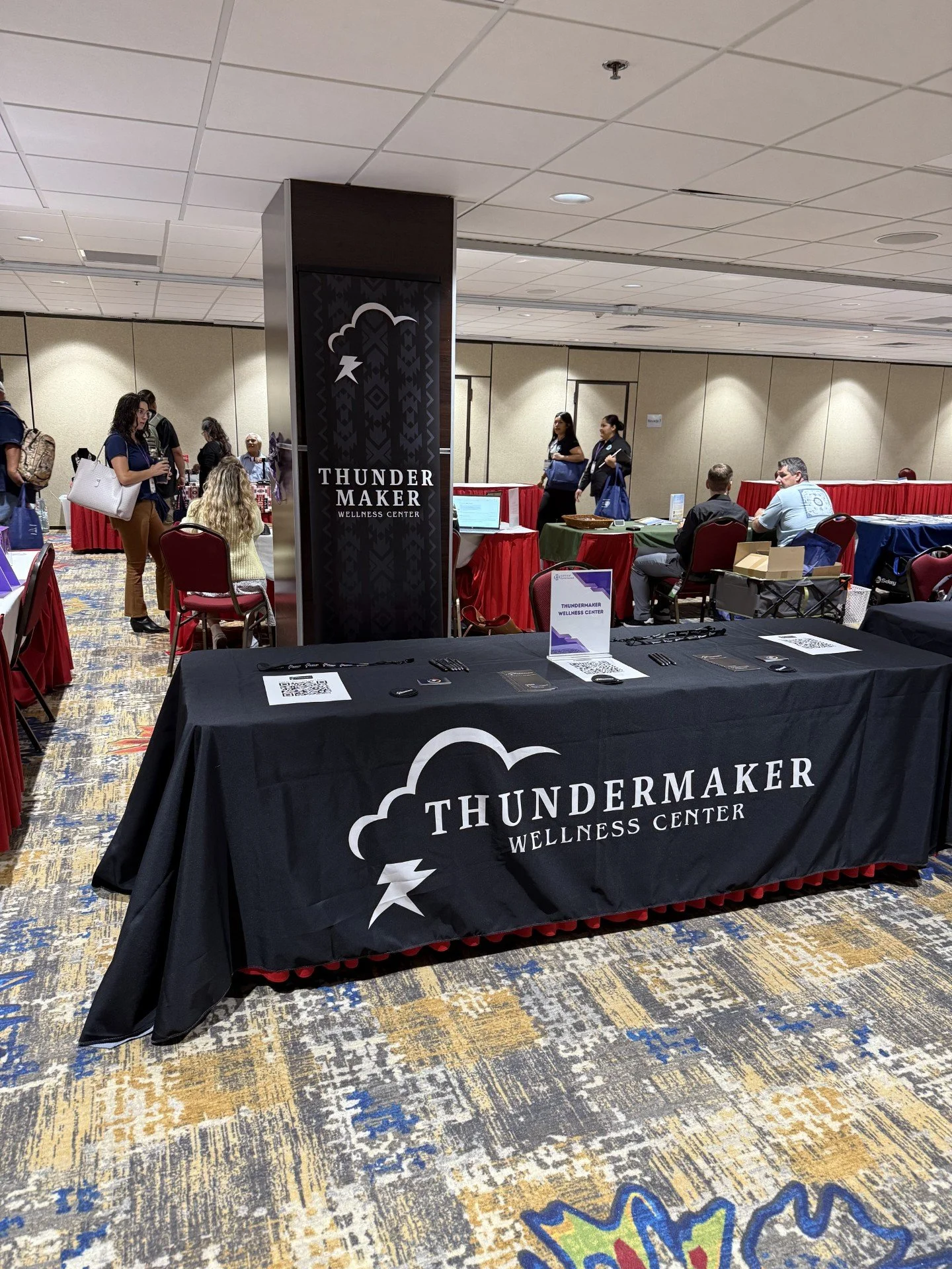 Exhibition booth at the Washoe Tribal Opioid Conference with a black tablecloth displaying the logo and name of ThunderMaker Wellness Center at an indoor event, with people walking and talking in the background.