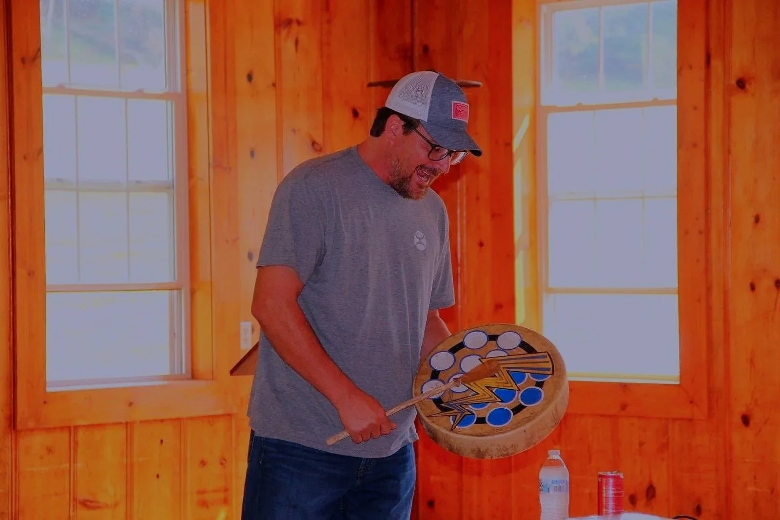 A man with a beard and glasses, wearing a gray t-shirt and a baseball cap, is playing a game of cornhole in a wooden room with large windows.