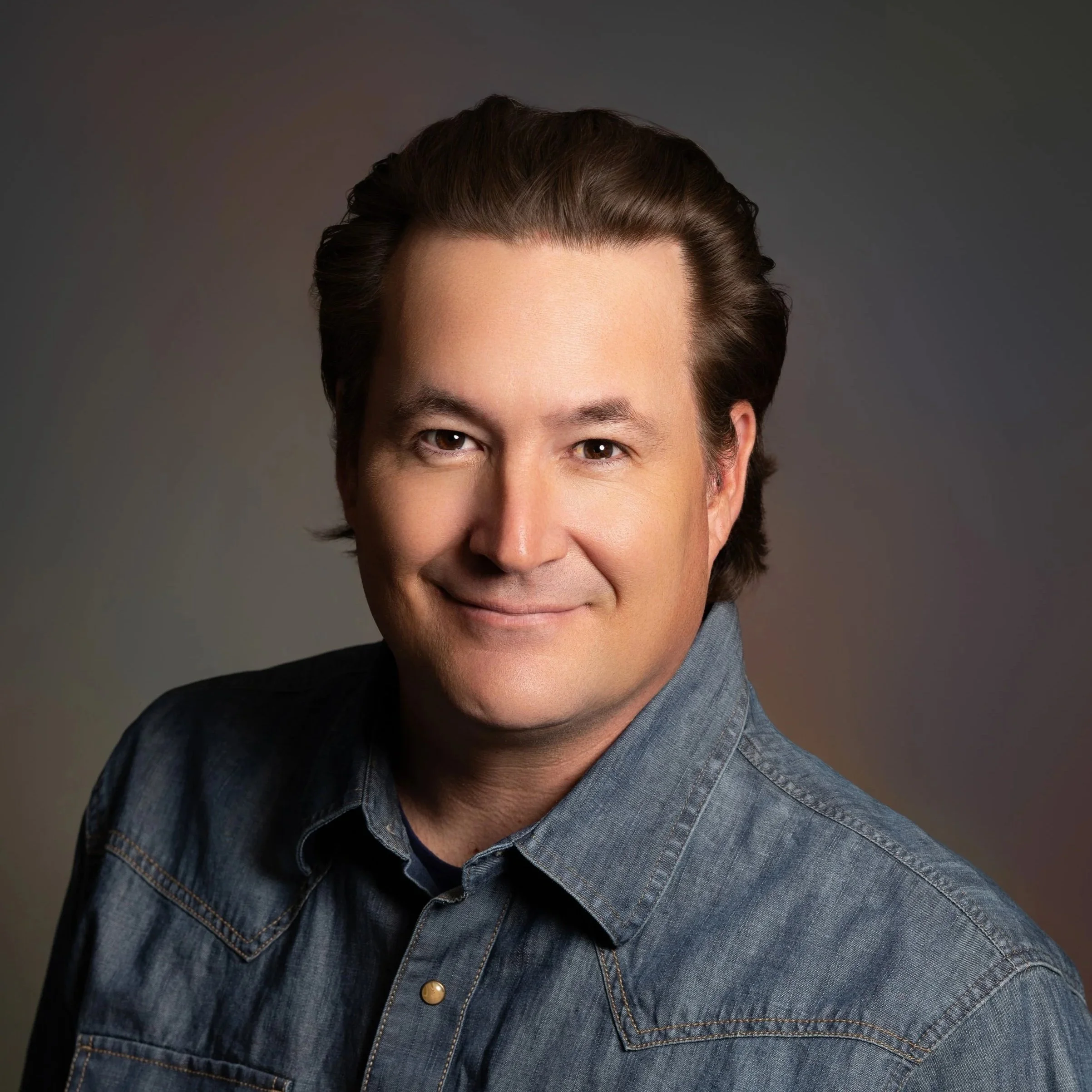 Arrow Funmaker, Founder and Chief Cultural Integration Officer of Thundermaker Wellness in a professional headshot wearing a demin button up shirt on a dark grey background. 