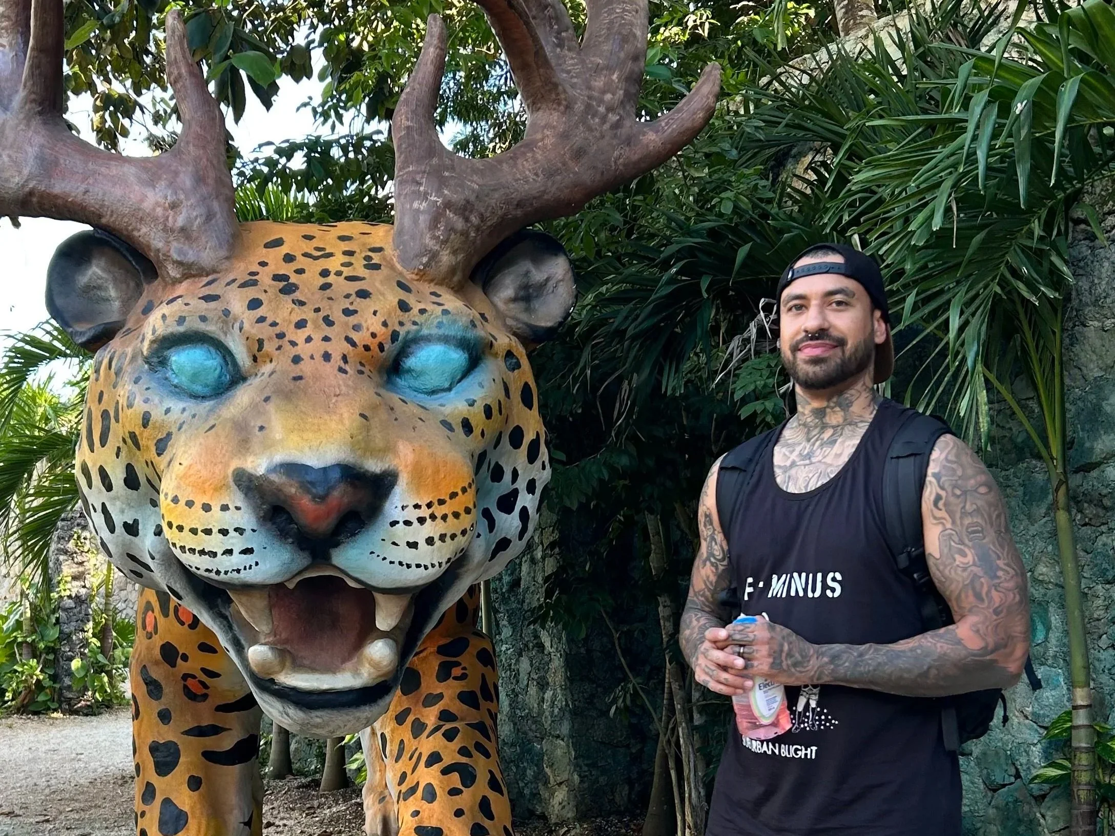 A man with tattoos wearing a black tank top and a backward cap, standing next to a large, colorful animal statue with antlers, resembling a jaguar or leopard with blue eyes, set in a lush, green outdoor environment.
