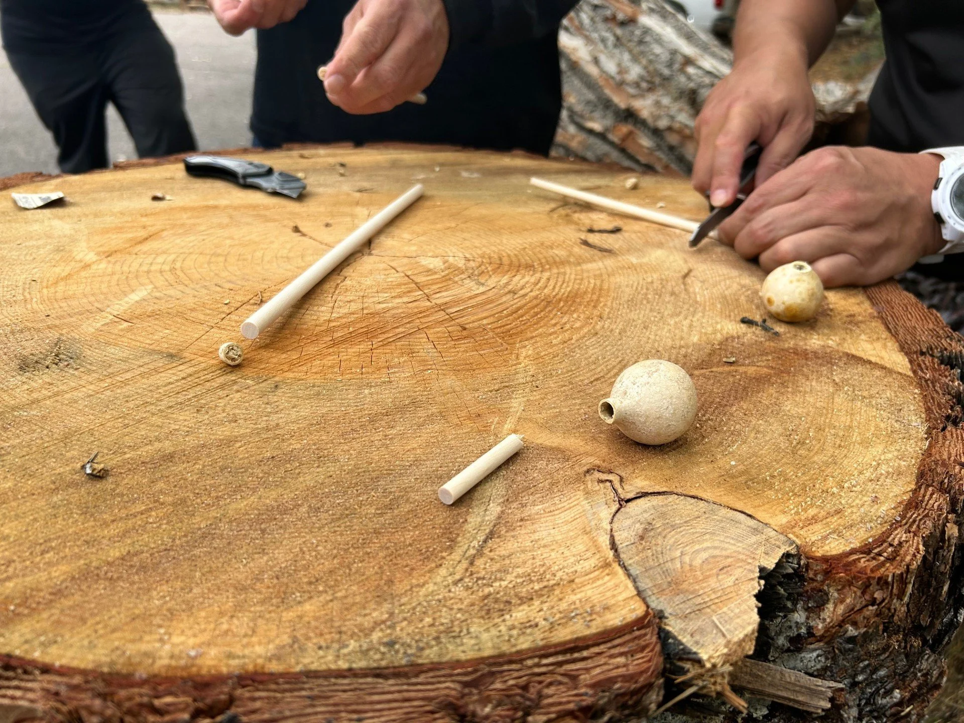 A hand is holding a small beaded bracelet with black, white, and green beads, while a wooden loom and other craft supplies are visible on a wooden surface.