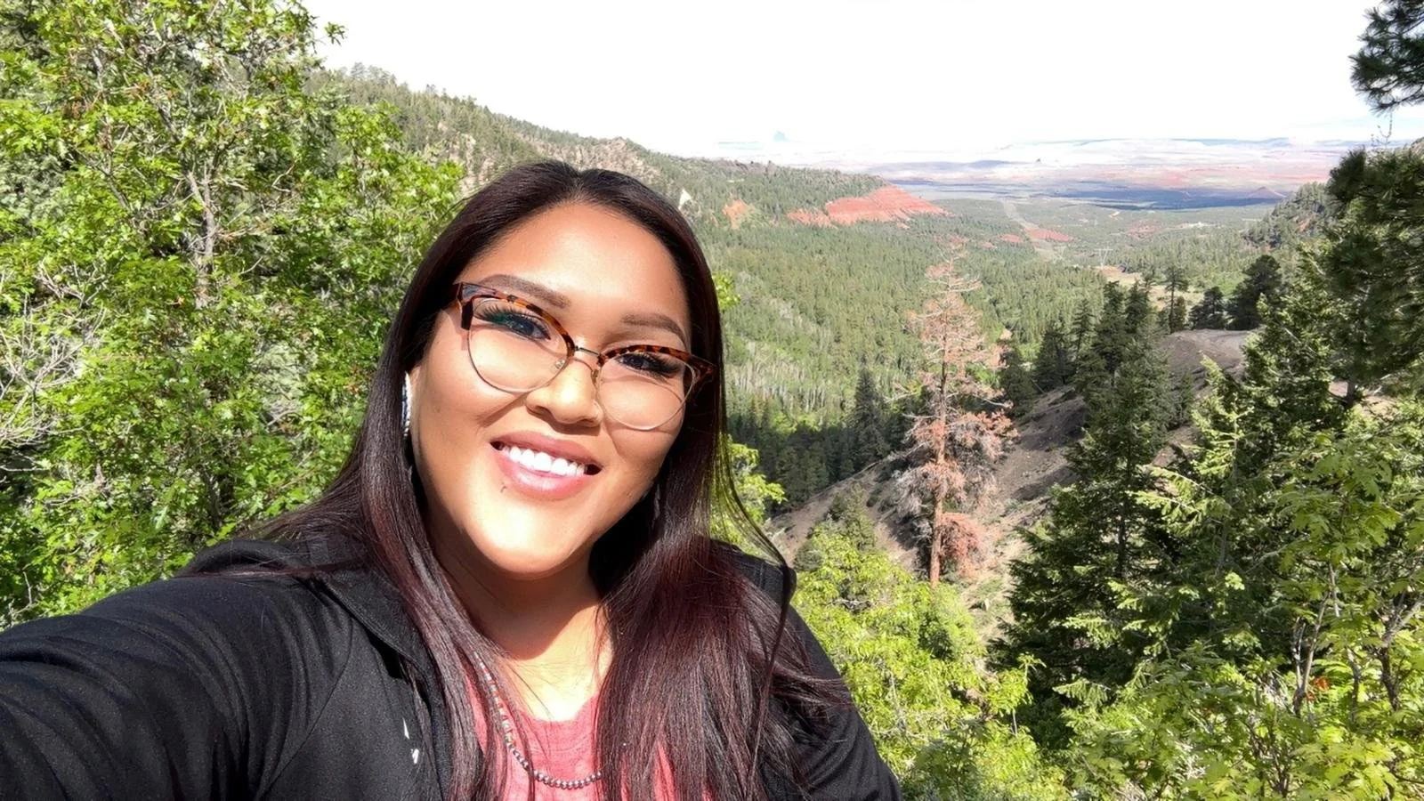 A woman with glasses and long dark hair smiling in front of a scenic mountain landscape with trees and distant hills.
