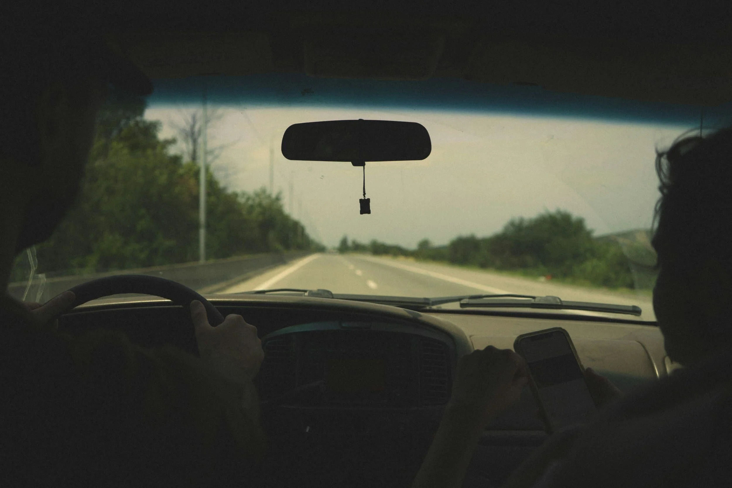 A picture of looking out of a windshield in a car driving on a highway with trees and bushes in the surrounding area