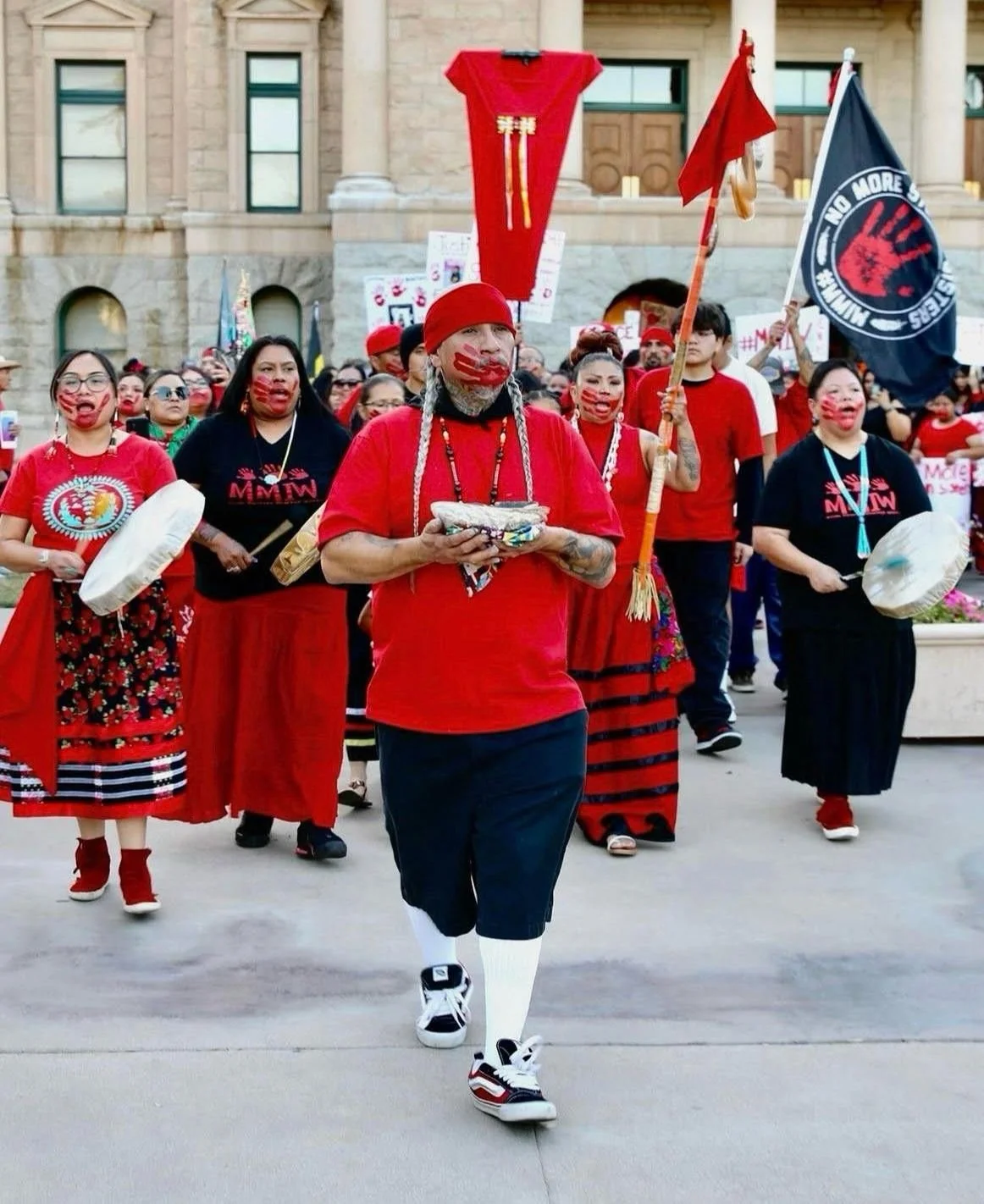 Group of Indigenous people participating in a protest or march, wearing red and black clothing, some holding flags and drums, with face paint and traditional accessories, in front of a government building.