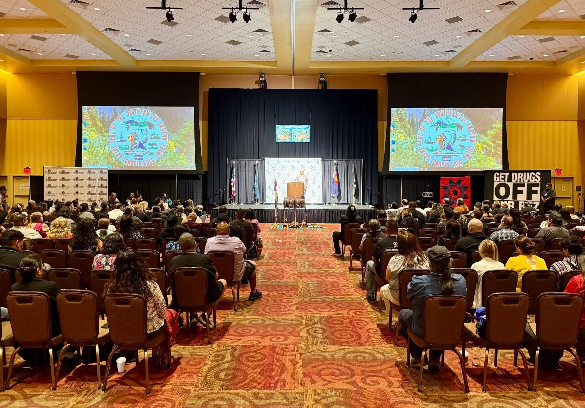 A large conference or rally in an auditorium with many attendees seated facing a stage. The stage has three flags, and two large screens display the logo of the Southern Ute Indian Tribe. There is a sign on the right side of the room that says 'GET DRUGS OFF OUR RES.'