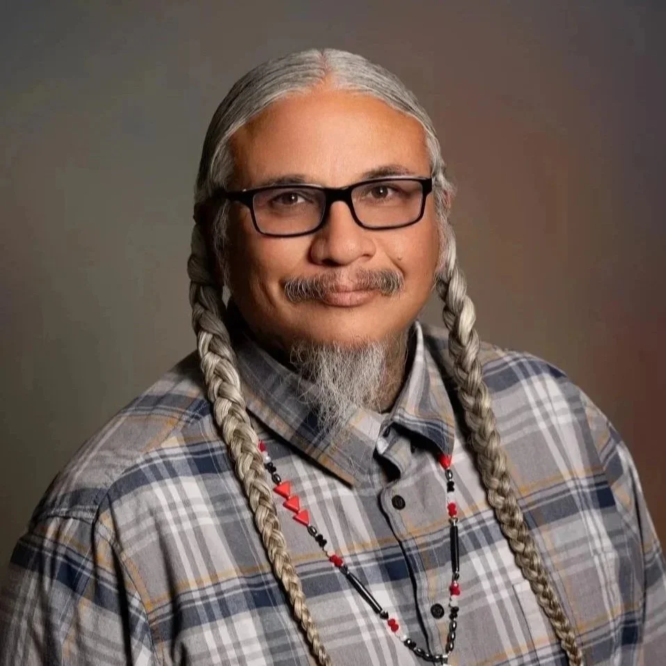 Solomon Acosta, Relative Support Specialist with Thundermaker Wellness, with long, gray braids, wearing glasses, a plaid shirt, and a necklace with beads, smiling against a plain background.
