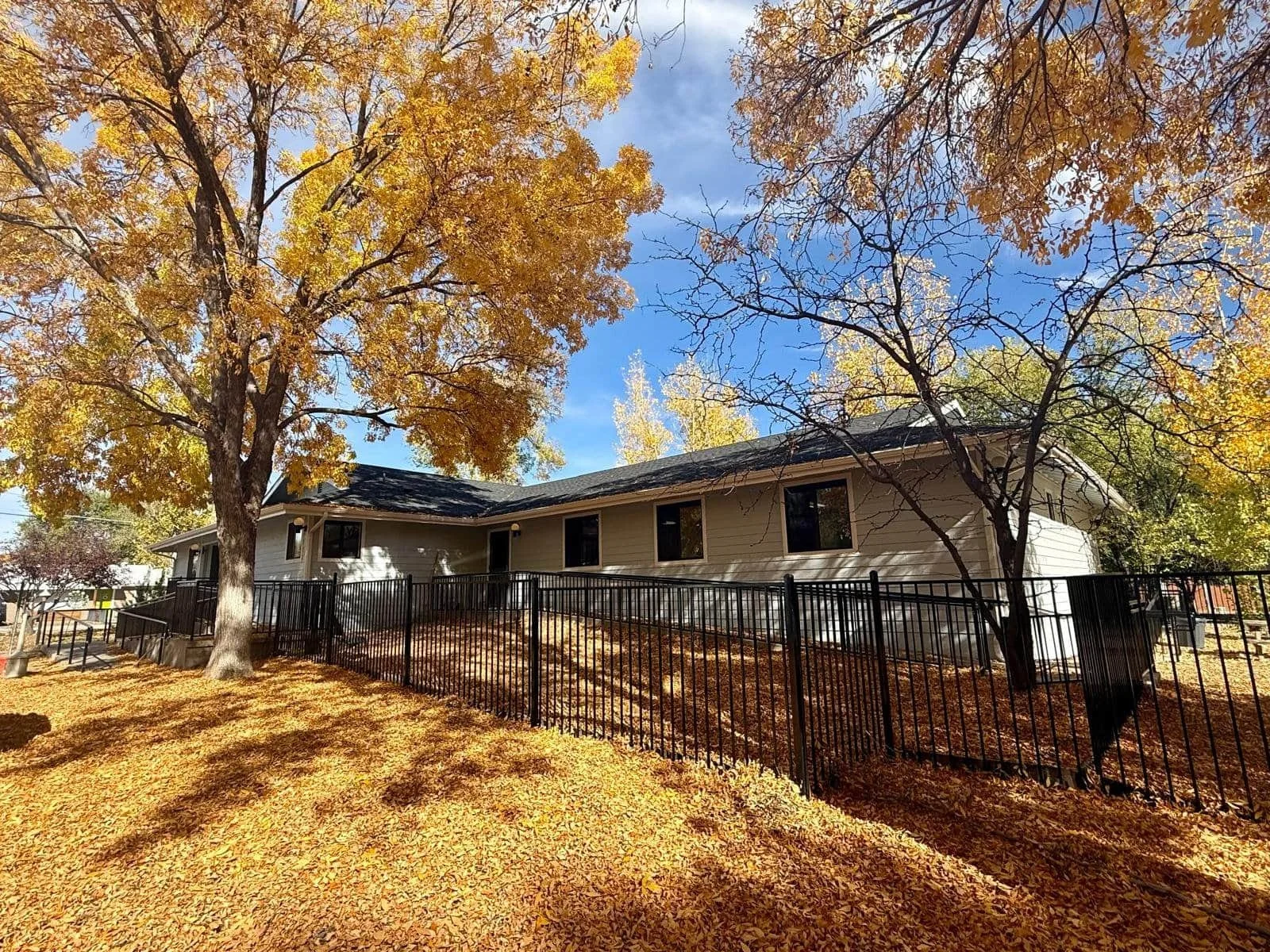 A blue outpatient substance use and mental health office with black windows and roof, surrounded by trees with bright yellow autumn leaves, a black metal fence, and a yard covered with fallen leaves, under a partly cloudy blue sky.