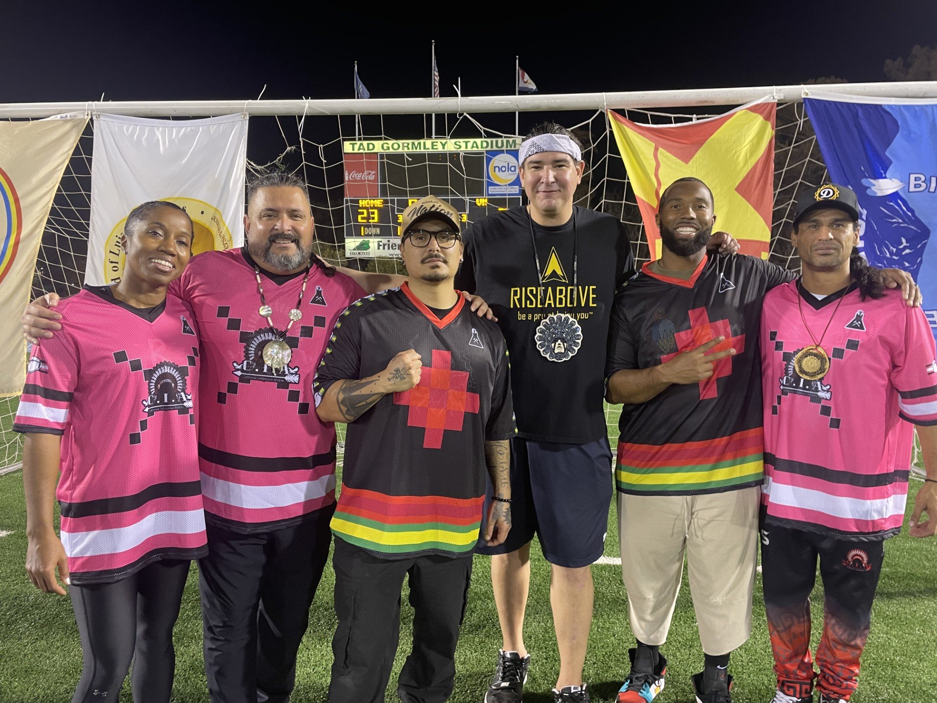 Group of six indigenous celebrities standing on a soccer field at night, wearing matching athletic jerseys for the Indigenous Girls Flag Football Team in New Orleans, LA