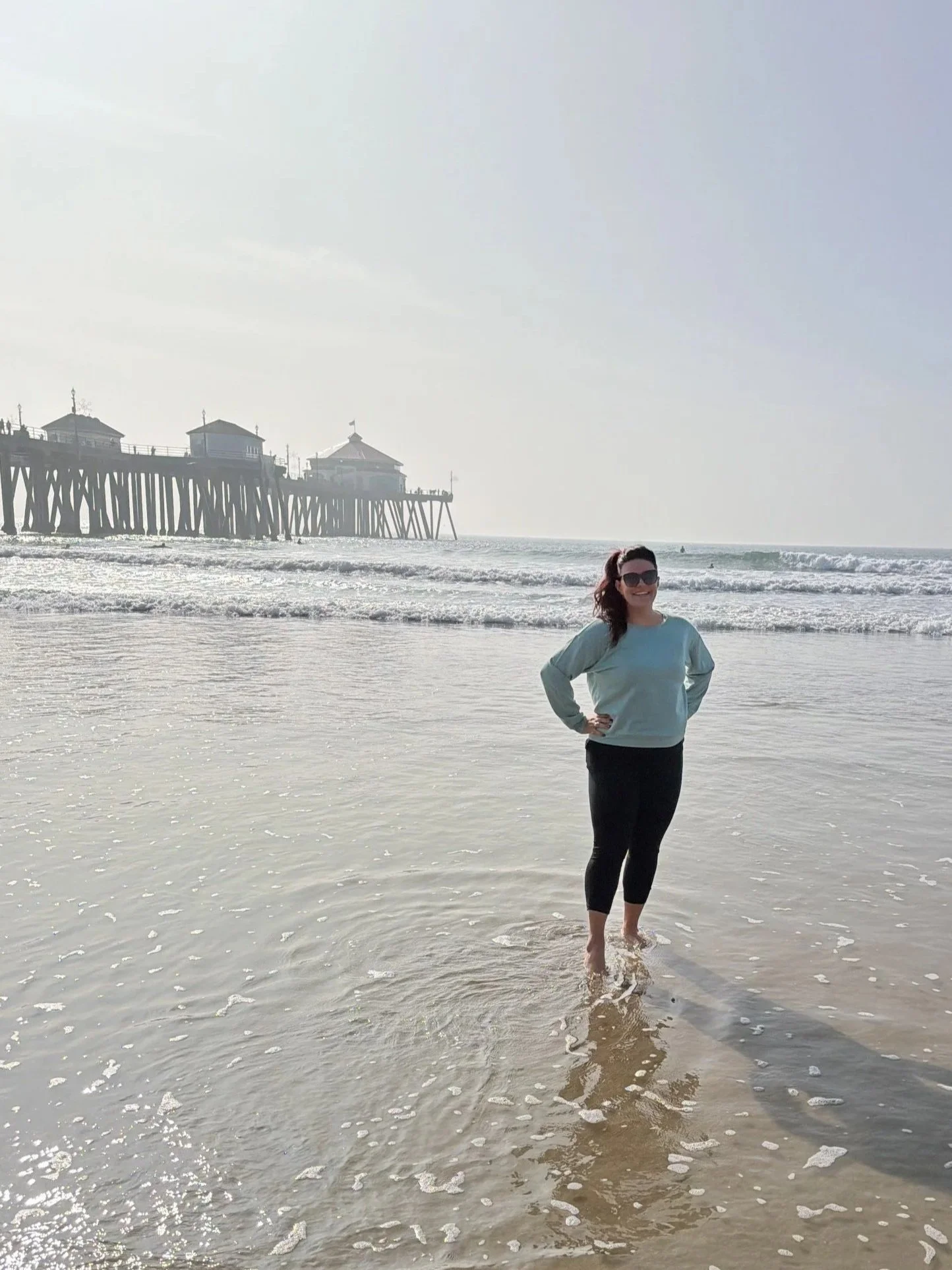 Bailey Gonzales, group facilitator at Thundermaker Wellness, walking in shallow ocean water near a beach with a pier and buildings in the background, smiling while wearing sunglasses and a light blue sweater.