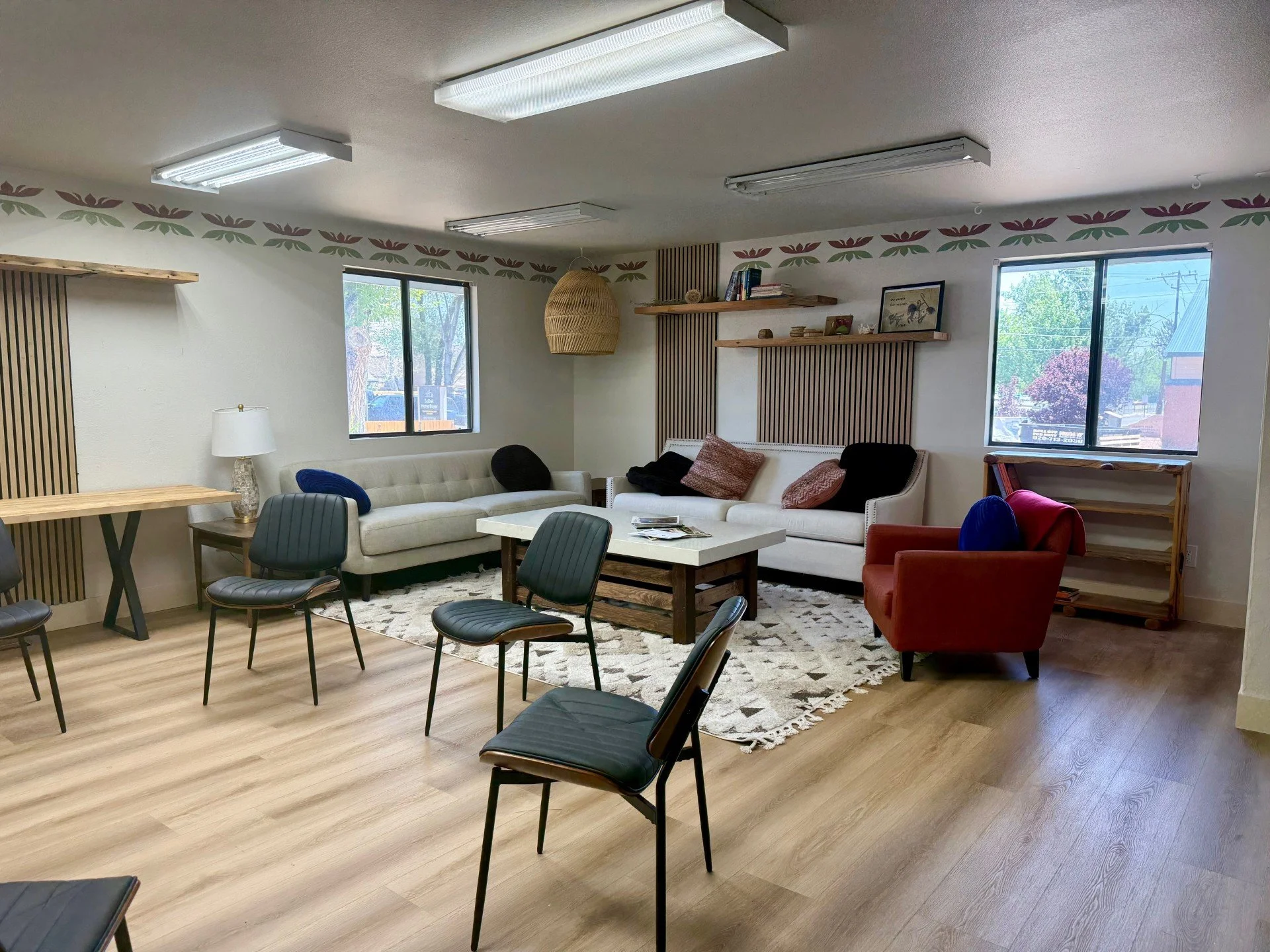 A cozy group therapy room with two white sofas, one red armchair, a wooden coffee table, and several black chairs. The room features wooden accents, a patterned rug, and natural light from two windows. Decorations include indigenous art pieces.