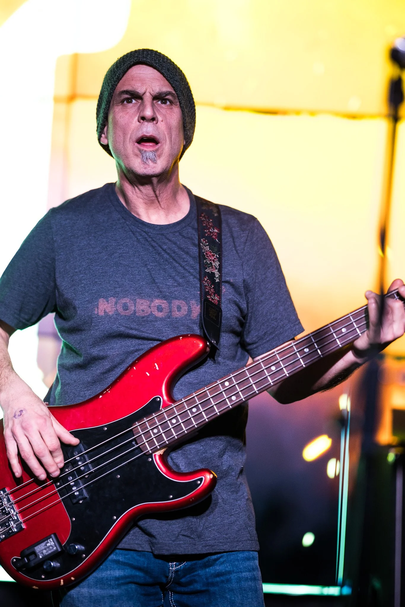 A man wearing a gray t-shirt and a knit beanie playing a red electric bass guitar on stage.