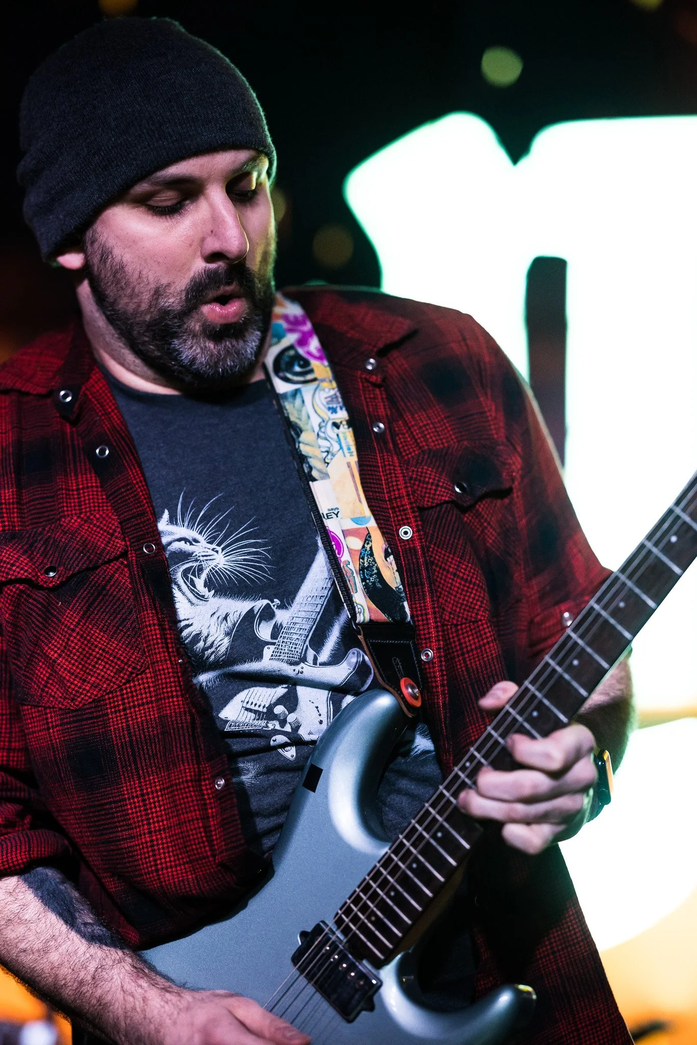 Man with a beard playing an electric guitar on stage, wearing a black beanie, a black T-shirt with a tiger and guitar graphic, and a red plaid shirt.