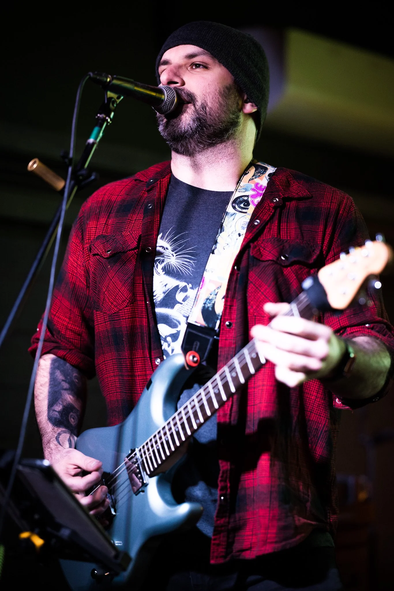 A musician with a beard and tattoos wearing a black beanie, red plaid shirt, and black graphic T-shirt, playing an electric guitar and singing into a microphone on stage.