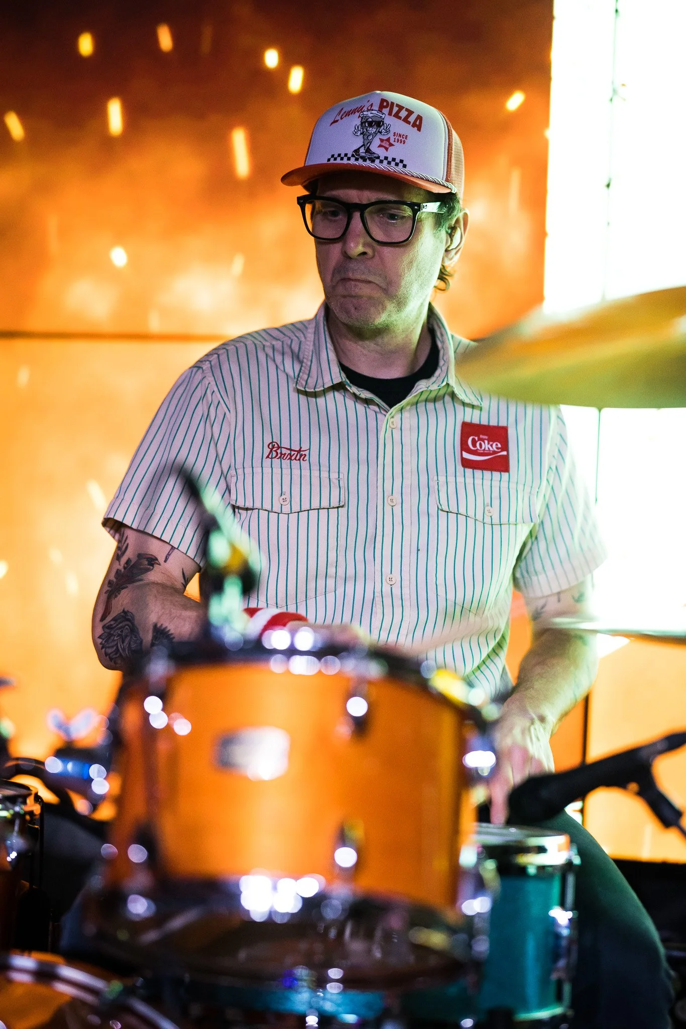 A man wearing glasses, a striped shirt with Chuck E. Cheese and Coke patches, and a baseball cap with a pizza theme, playing drums during a performance.