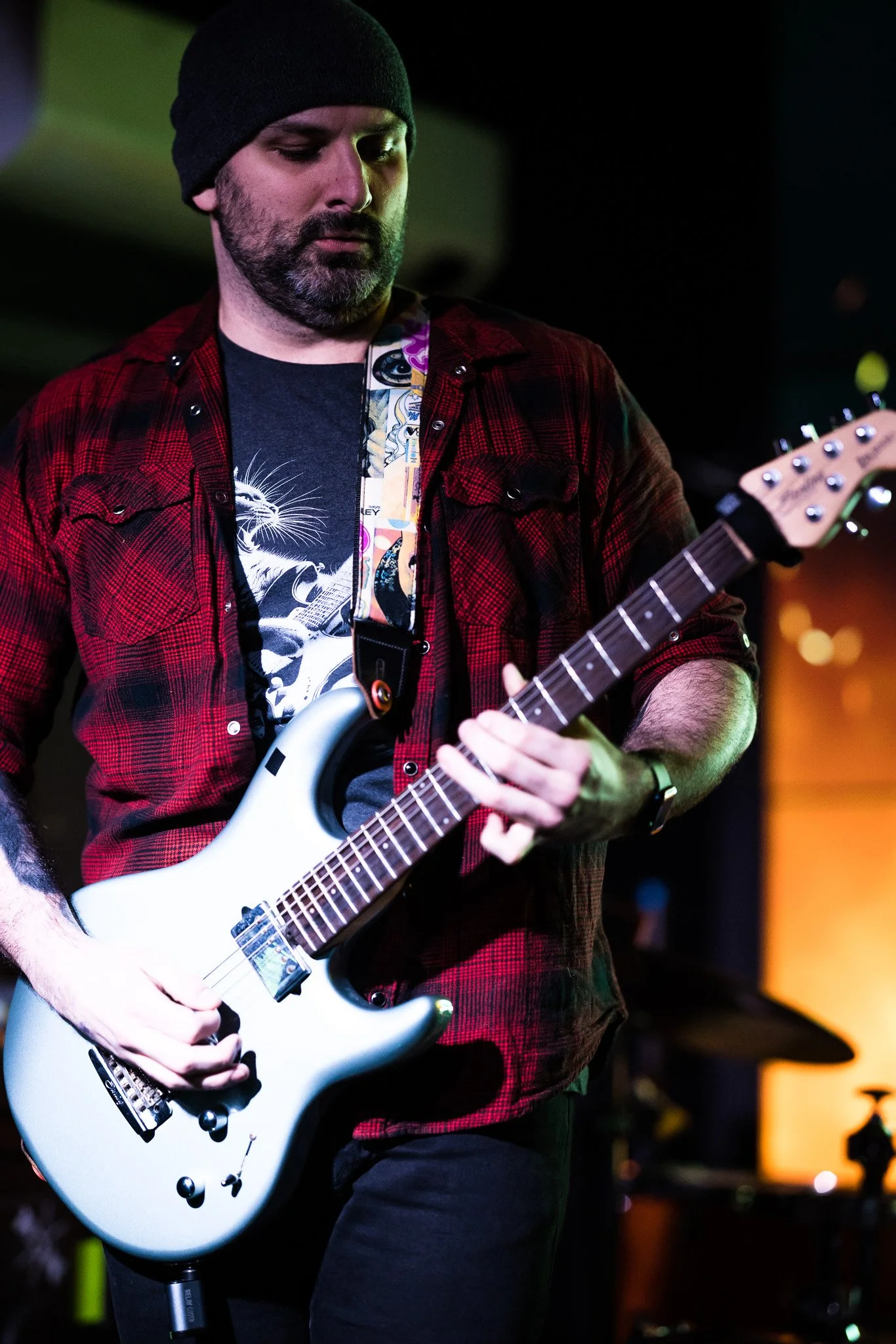 A man playing an electric guitar on stage, wearing a black beanie, a red and black plaid shirt, and a graphic T-shirt, with a colorful guitar strap.