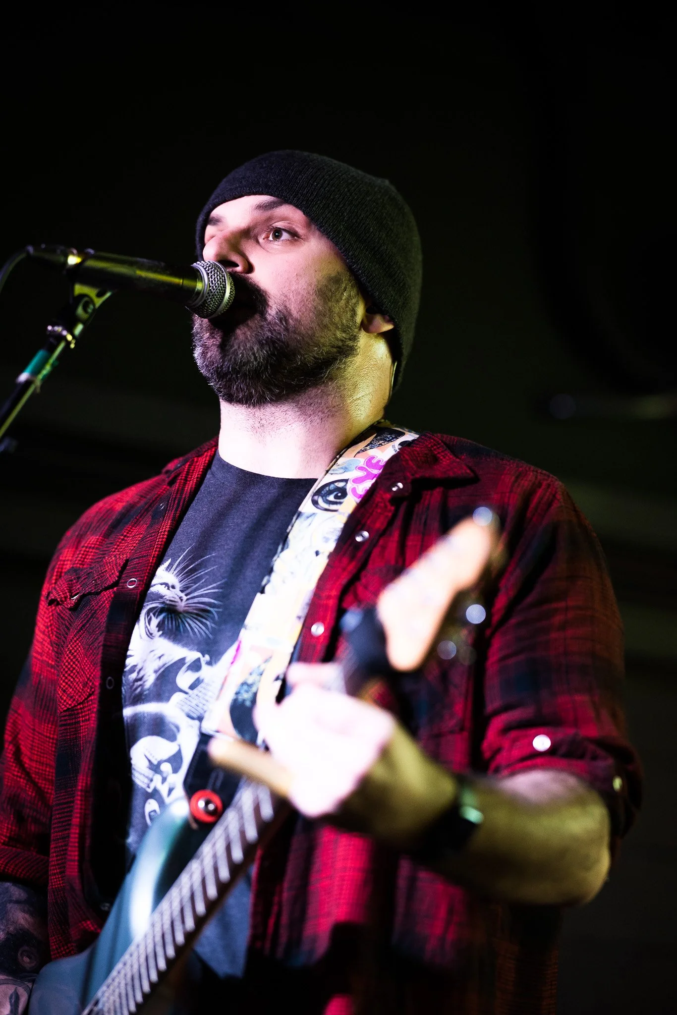 A man with a beard wearing a black beanie and red plaid shirt, playing an electric guitar and singing into a microphone on stage.