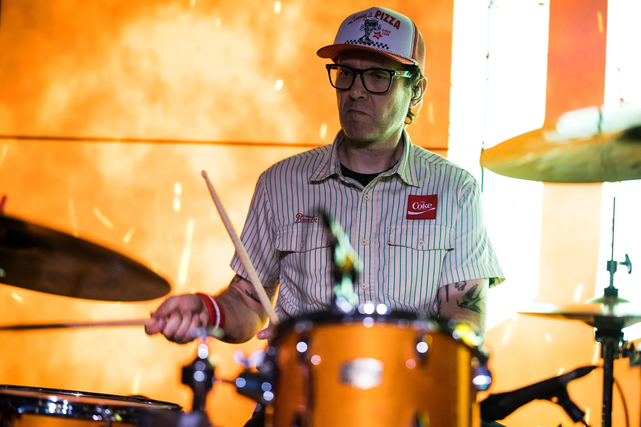 A man wearing glasses, a striped shirt with patches, and a cap with a pizza logo, playing drums against an orange background.