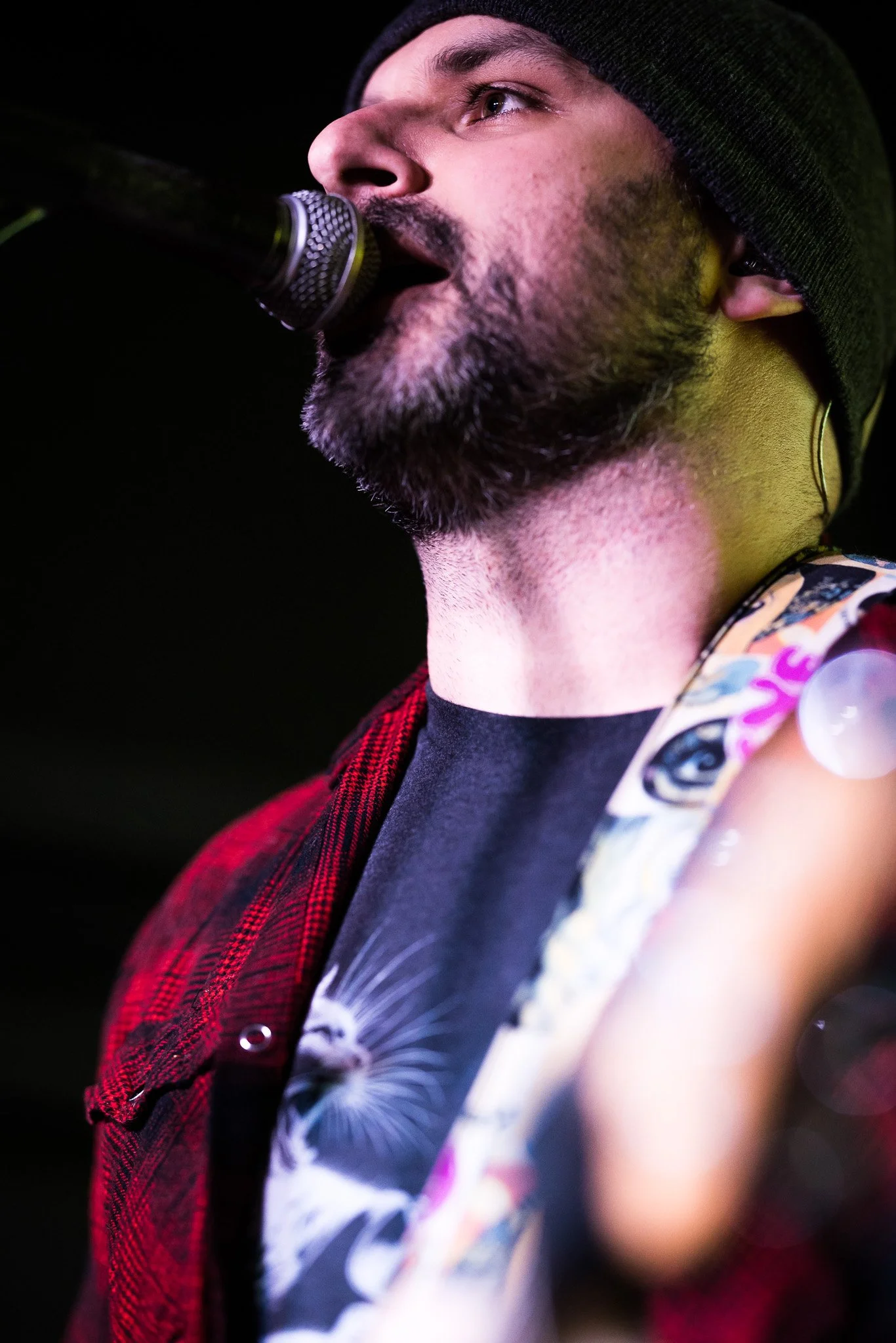 Close-up of a singer performing on stage, wearing a black beanie, with a microphone near his mouth, and sporting a beard, with details on his face and part of his colorful clothing visible.