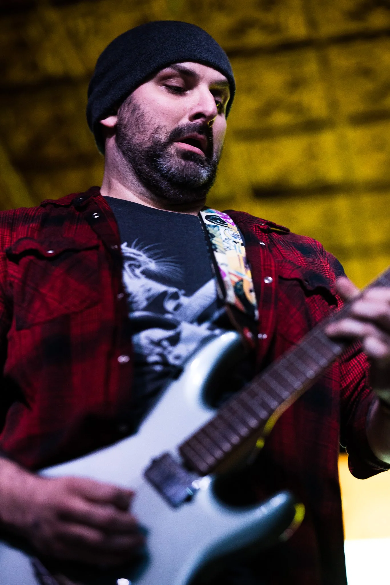 A man wearing a black beanie, a red flannel shirt over a graphic t-shirt, is playing an electric guitar.