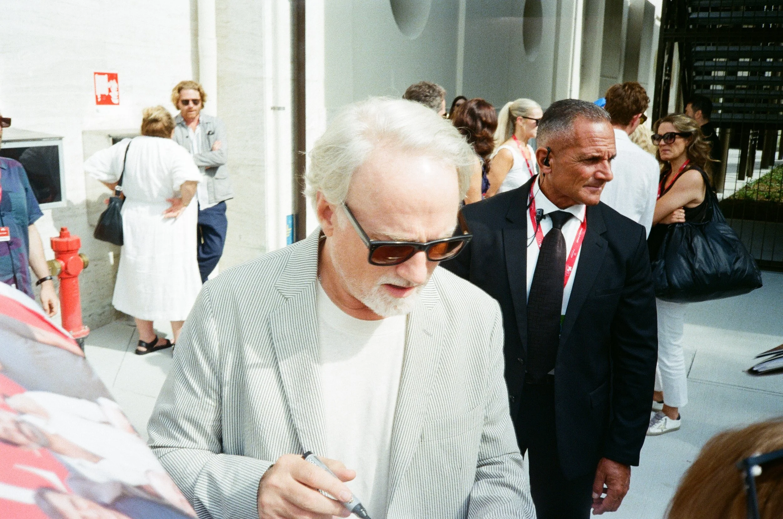 David Lynch firma un autografo, indossa una giacca a righe e una maglietta chiara. Dietro di lui, un uomo in abito nero con una cravatta nera e un cordino rosso con badge, e altre persone in fila o in attesa.