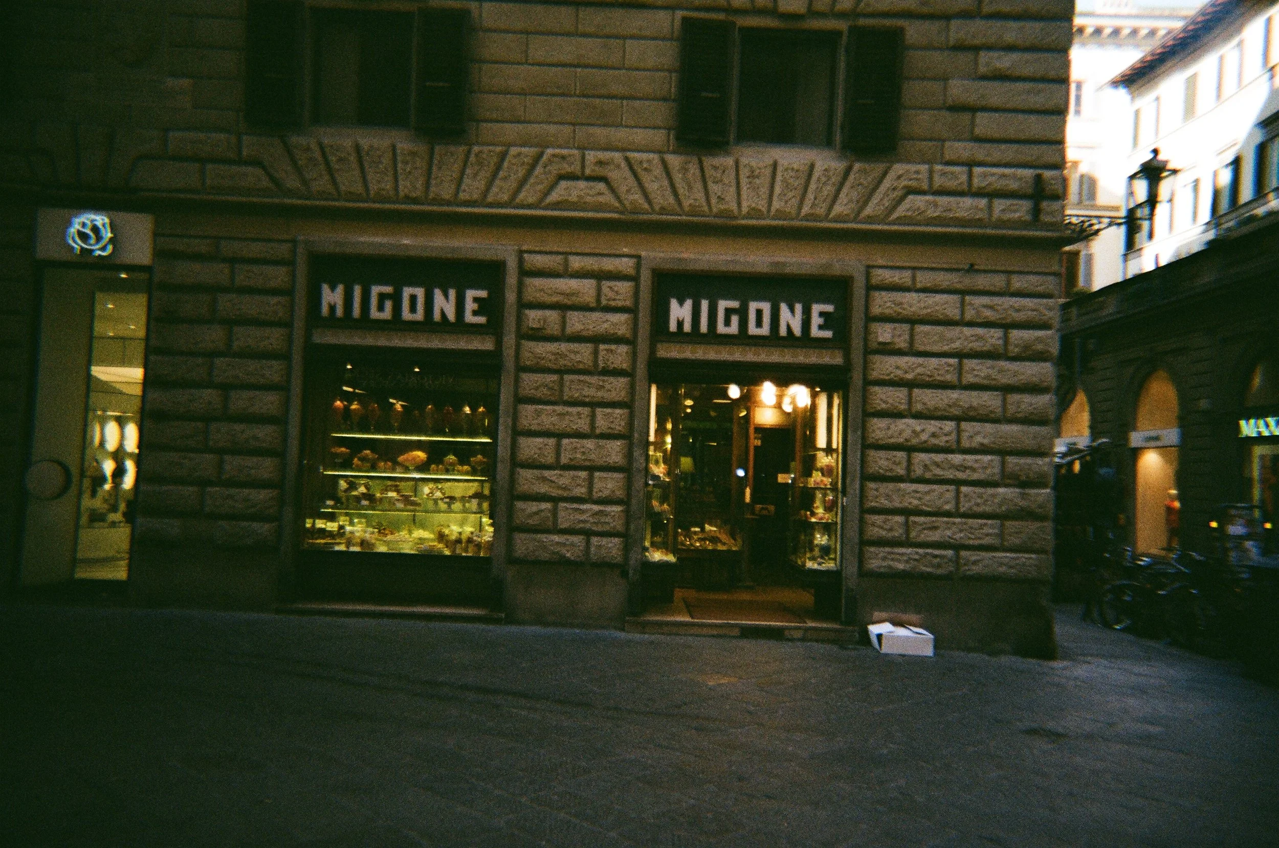 Vetrina di un negozio chiamato MIGONE, con luci che evidenziano prodotti in esposizione, situato in un edificio di pietra in un'area urbana.