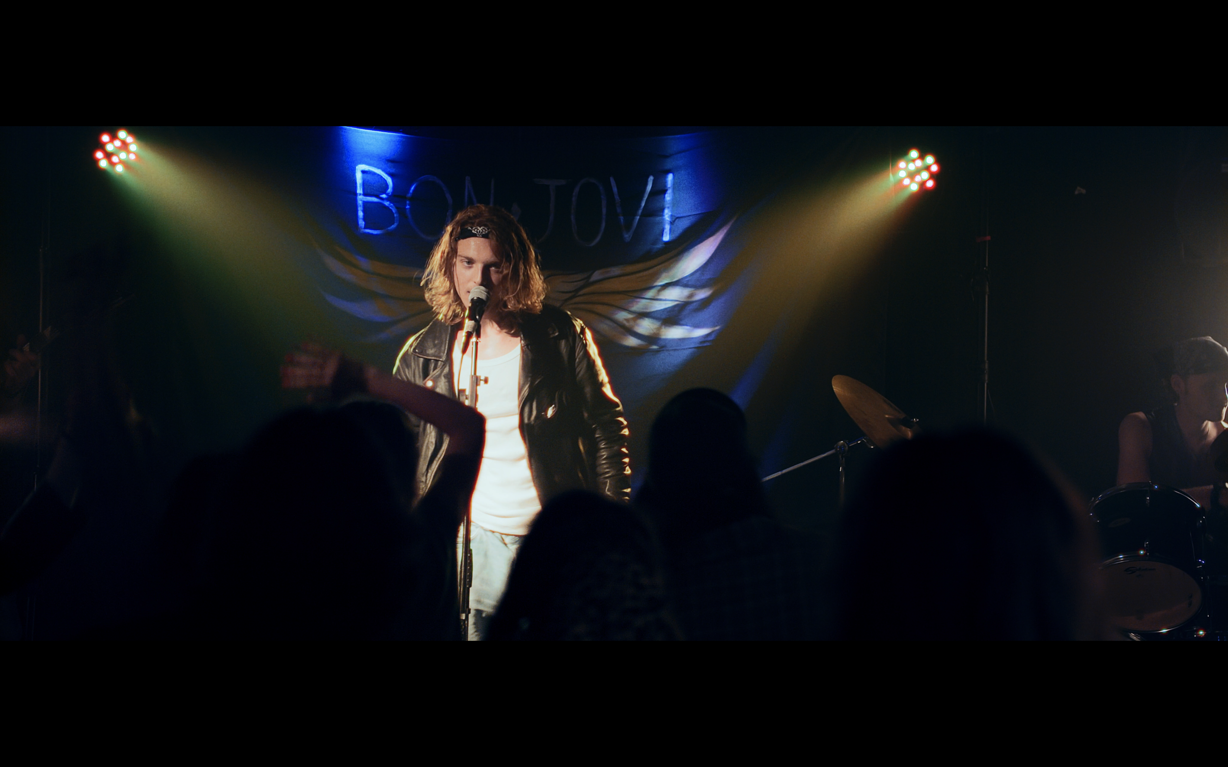 Artista con capelli mossi, giacca in pelle e microfono sul palco durante una performance musicale con luci colorate e pubblico in prima fila.