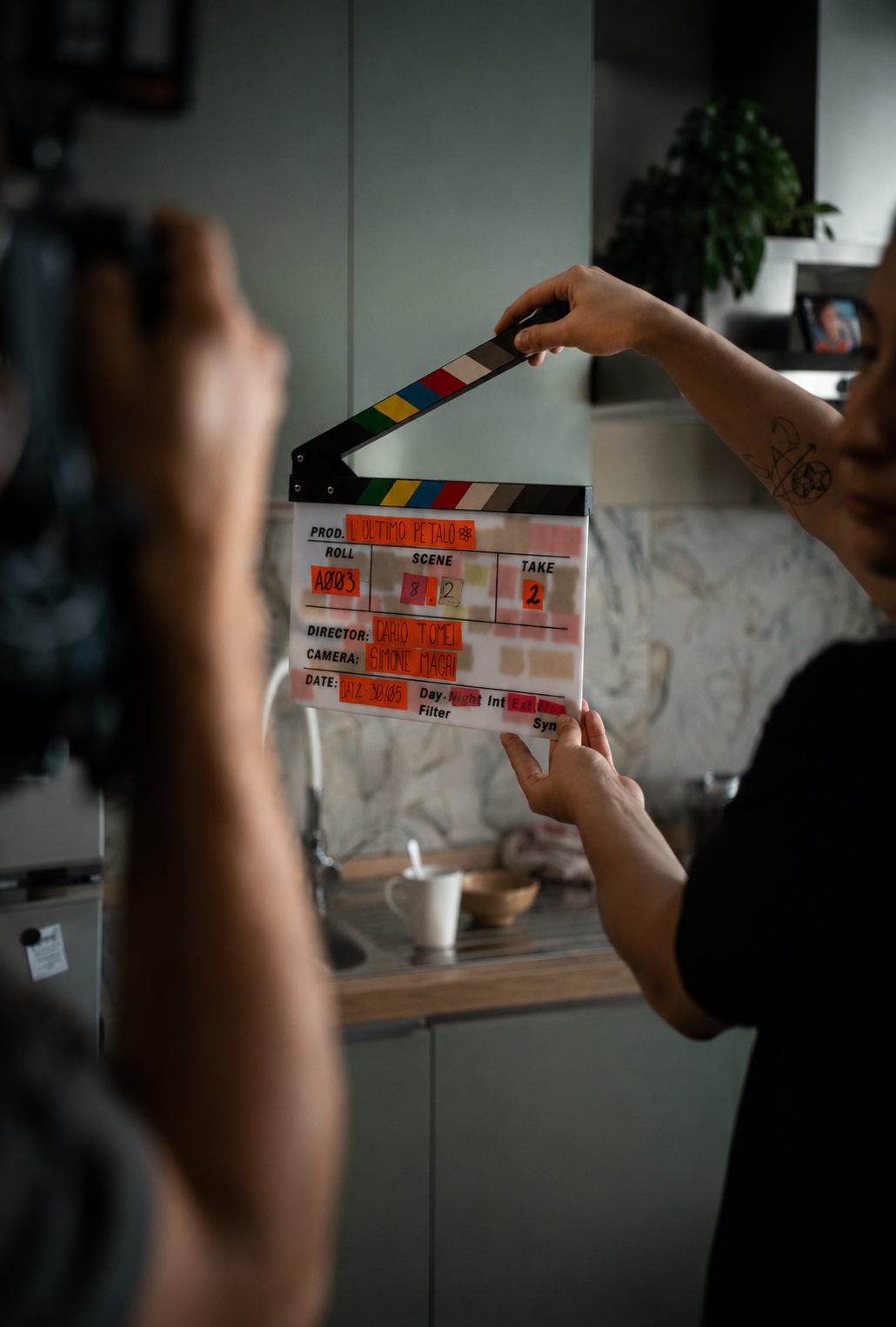 Immagine di una scena del set di un film con un clapboard, con persone che lavorano dietro le quinte, in un ambiente domestico con pareti di pietra e una pianta sullo sfondo.