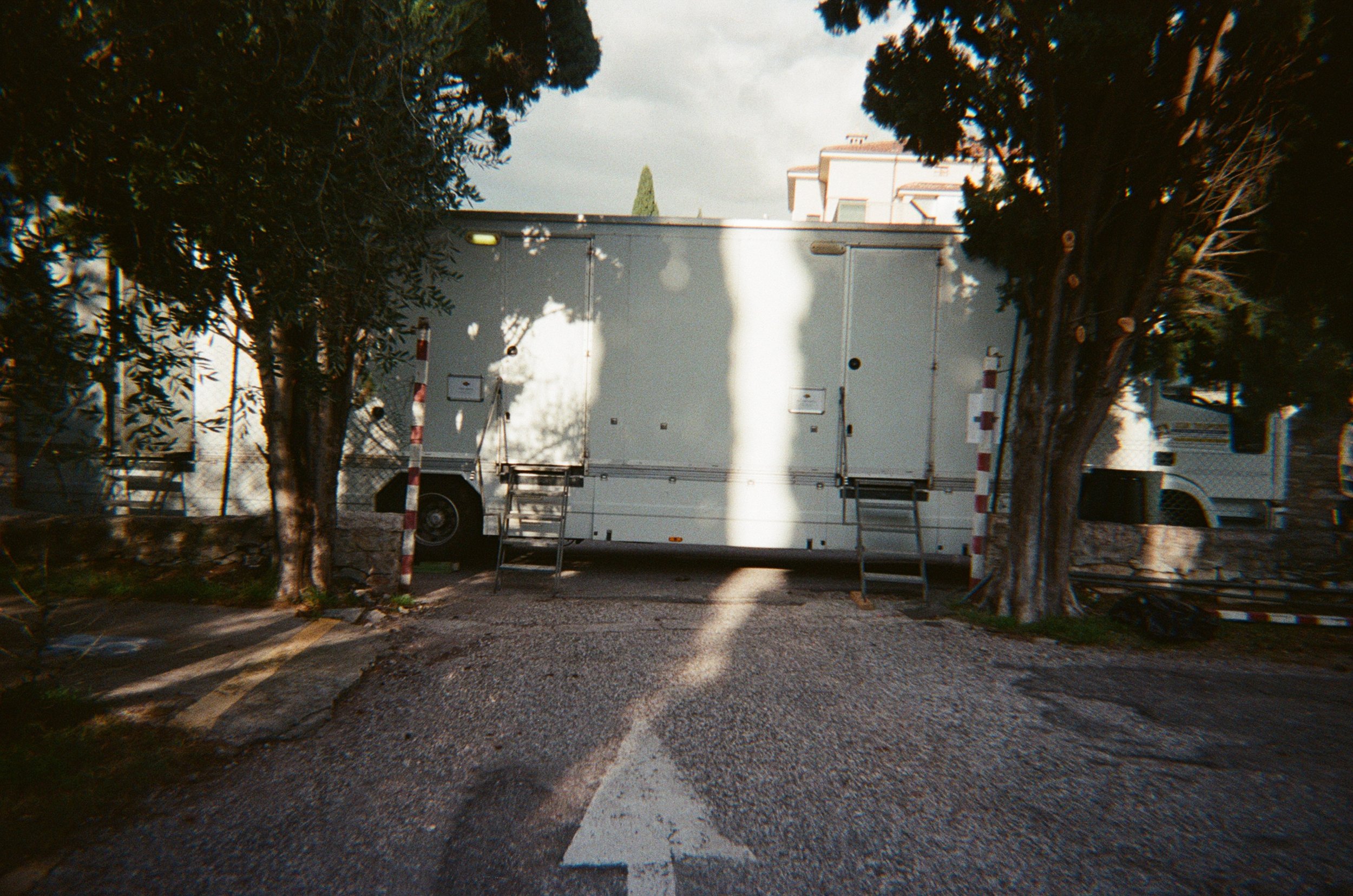 Vedo un camion bianco parcheggiato tra due alberi. In primo piano, una strada con una freccia bianca che indica avanti.