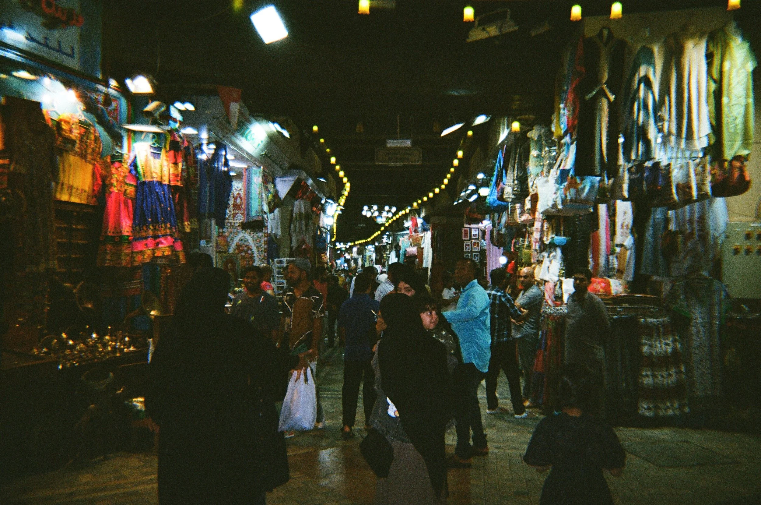 Veduta di un mercato coperto con bancarelle di abbigliamento e oggetti vari, affollato di persone che fanno shopping. L'ambiente è illuminato da luci calde e ci sono molti vestiti appesi e in vendita.